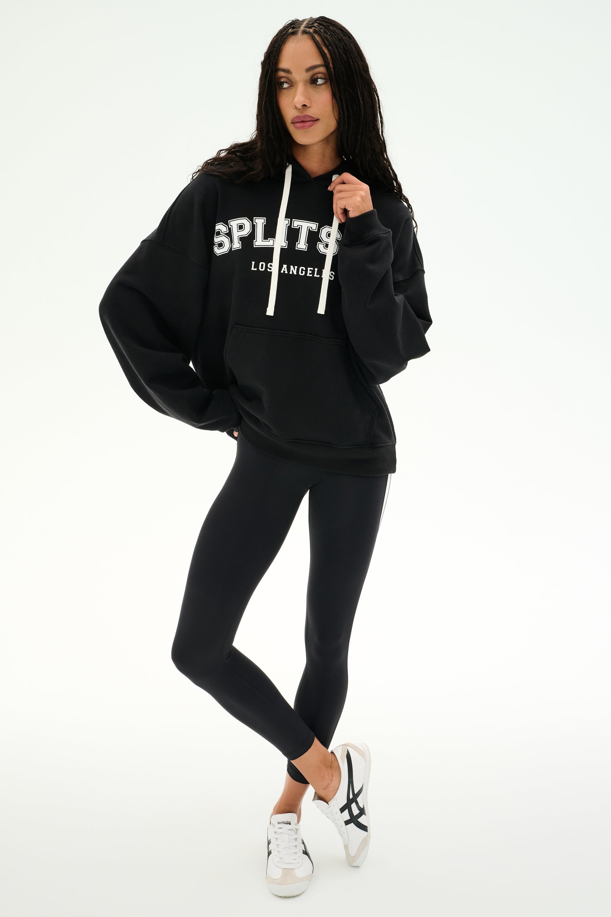 A person stands against a plain white background wearing the SPLITS59 Stella French Terry Oversized Hoodie in black, paired with black leggings and white sneakers.