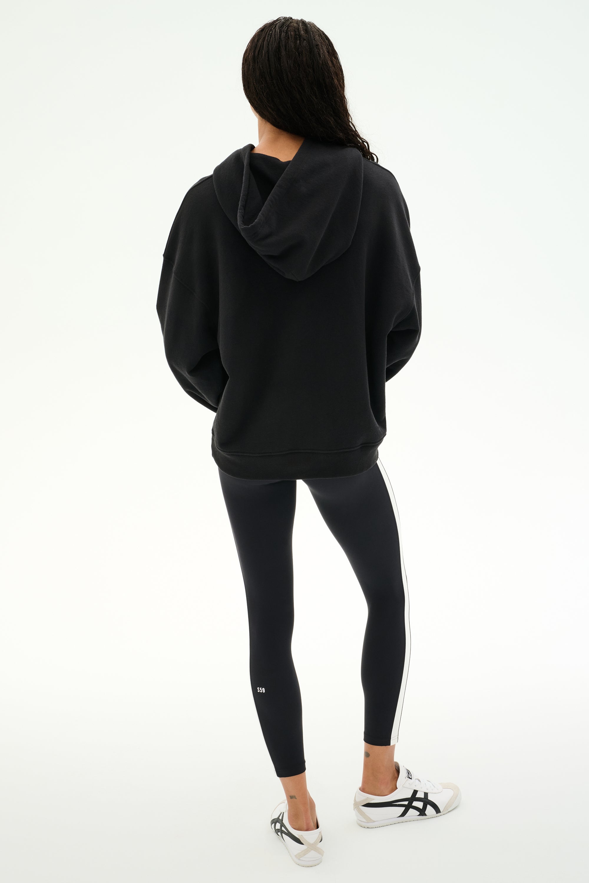 A person stands with their back to the camera, wearing the SPLITS59 Stella French Terry Oversized Hoodie in black, paired with black leggings featuring a white stripe and white sneakers, set against a plain white background.