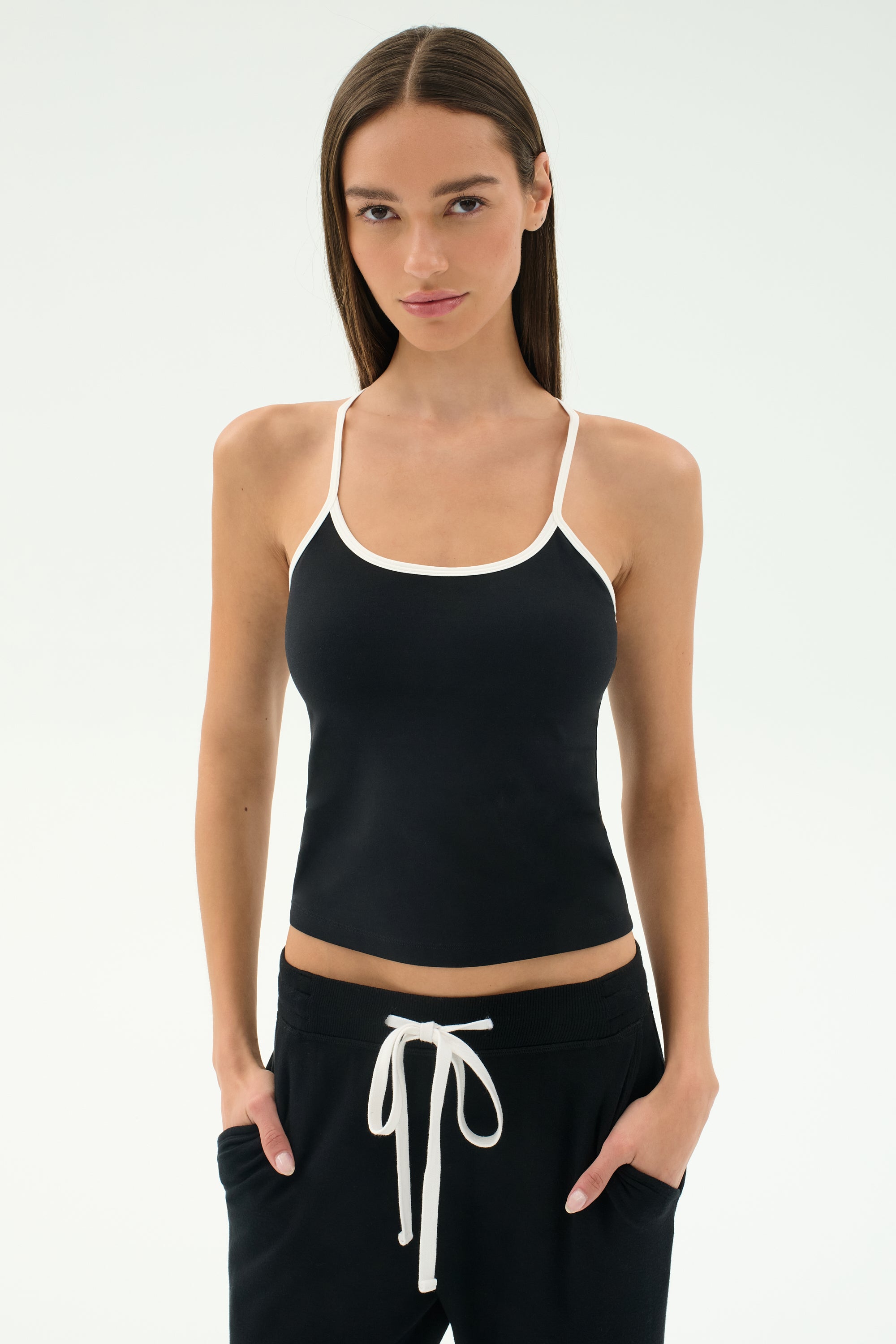 A woman stands facing forward in the SPLITS59 Airweight Tank - Black/White, paired with black pants with a white drawstring—an ideal Pilates outfit—hands in pockets, set against a plain light background.