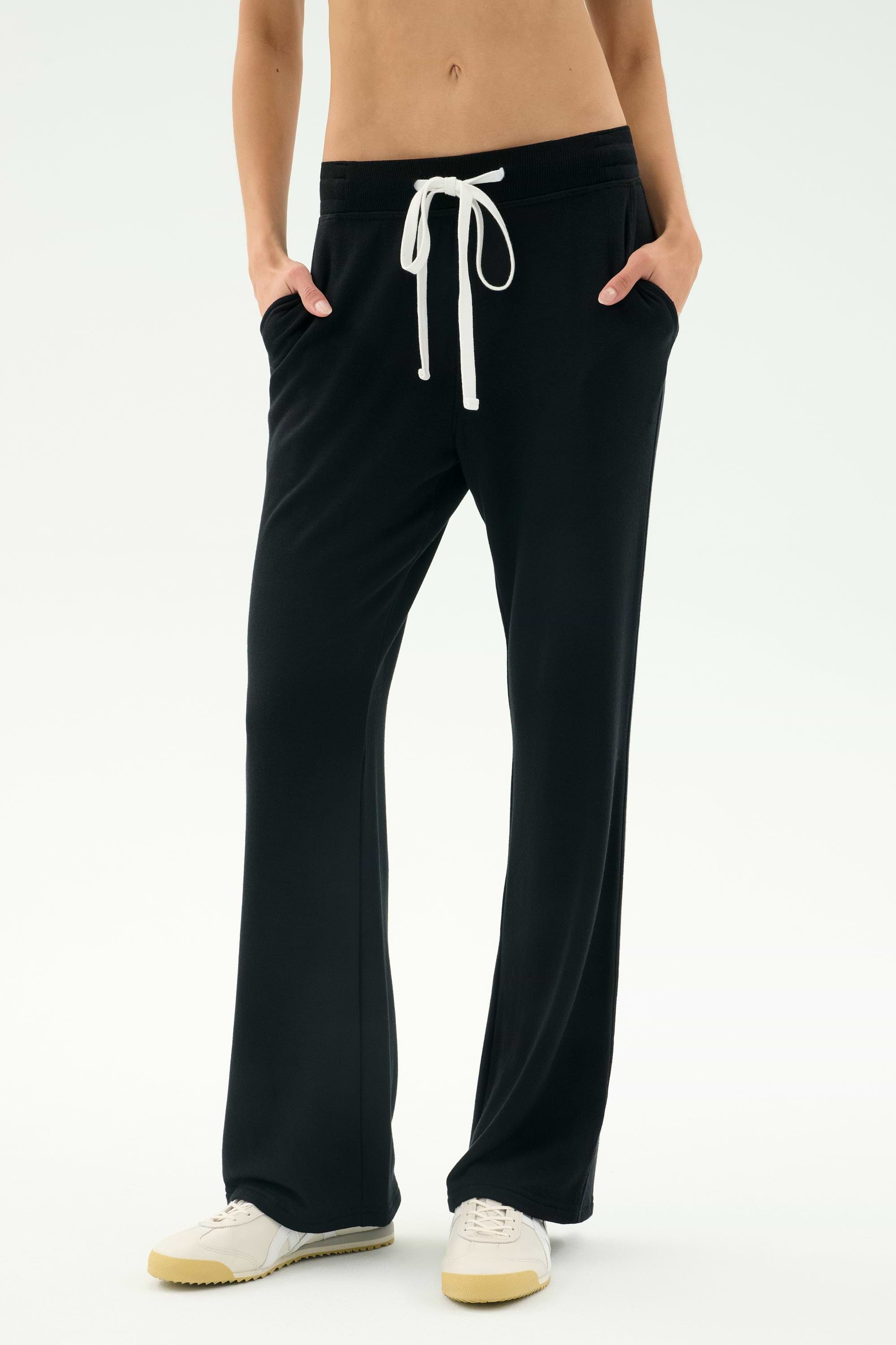 A person in SPLITS59 Raven Fleece Full Length Sweatpant - Black and white sneakers stands with hands in pockets against a plain background.