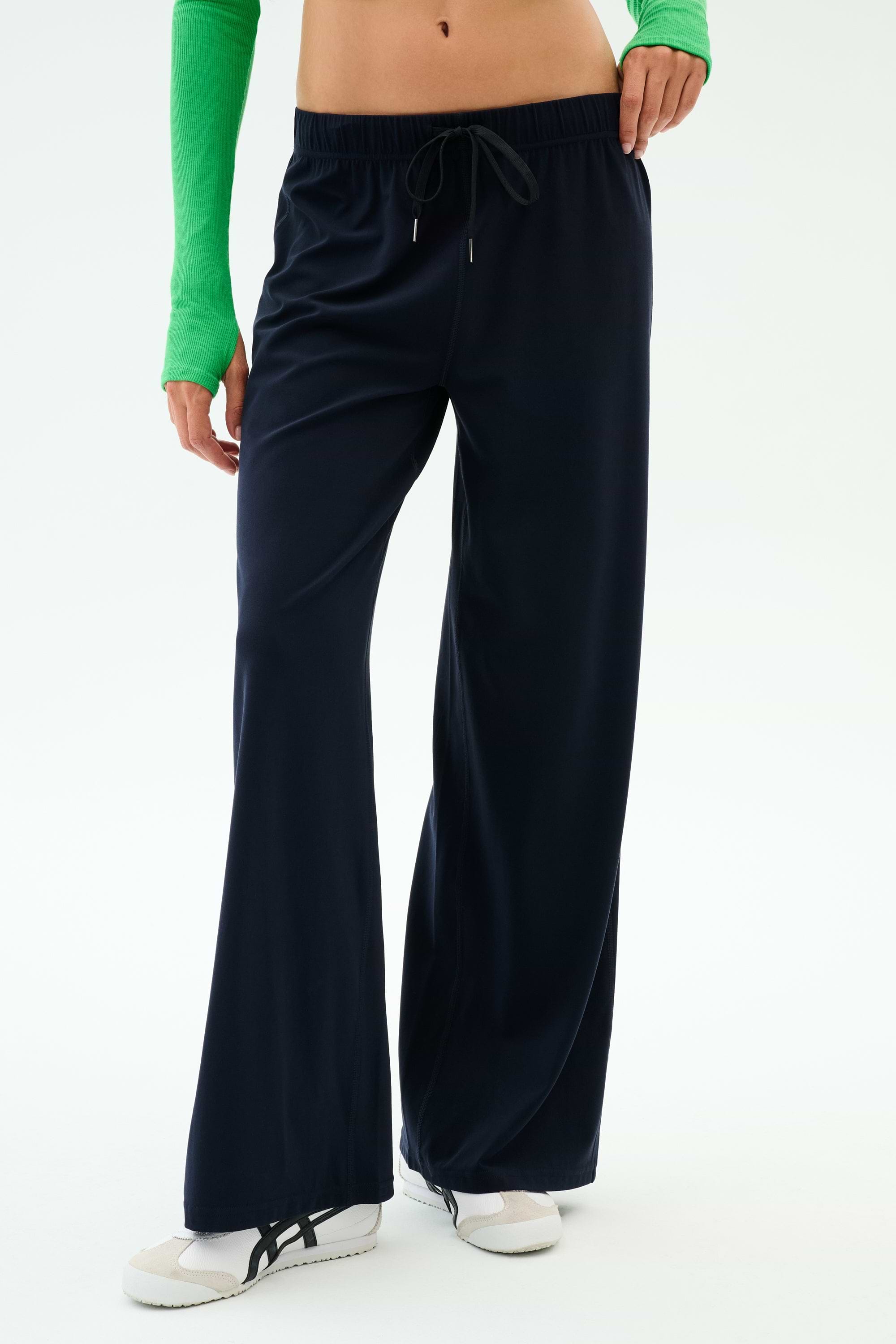 A person wears SPLITS59 Brynn Airweight Pant in Indigo, featuring a comfortable nylon-spandex blend, paired with a green long-sleeve top and white sneakers, standing against a plain white background.
