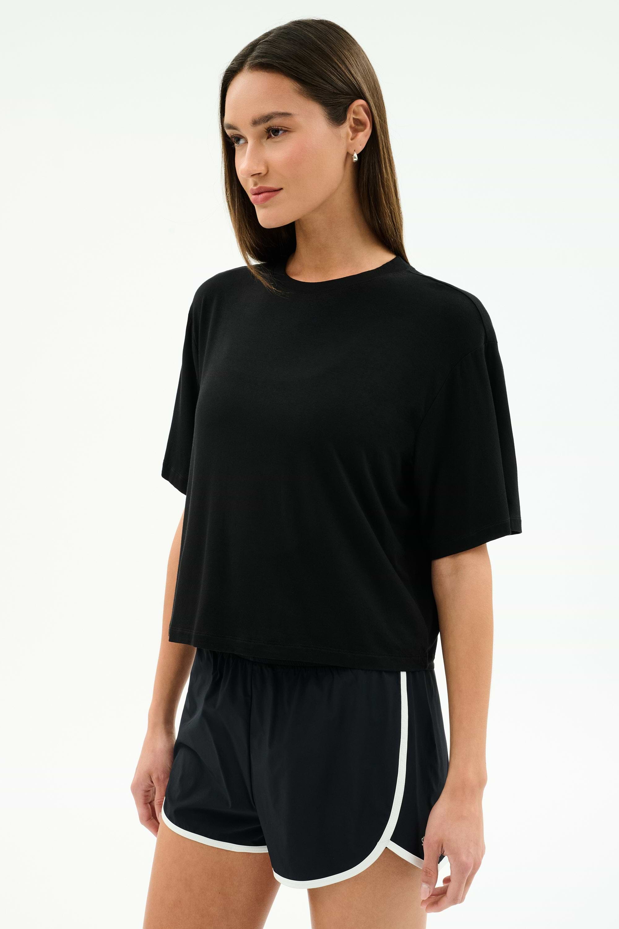 A woman wears the SPLITS59 Djuna Oversized Jersey Tee in black, paired with black athletic shorts with white trim, standing against a plain light background.