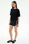 A woman stands against a plain background wearing a black spandex t-shirt, SPLITS59 Megan Woven Nylon Run Short in Black/Creme, and black Adidas sneakers.