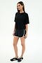 A woman stands against a plain background wearing a black spandex t-shirt, SPLITS59 Megan Woven Nylon Run Short in Black/Creme, and black Adidas sneakers.