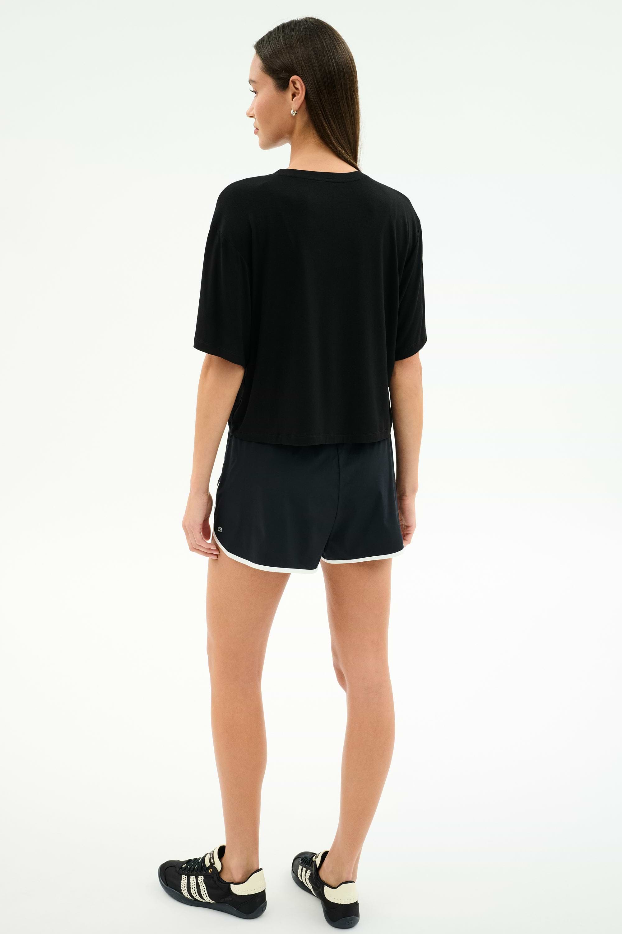 A woman faces away, wearing a loose black tee and SPLITS59 Megan Woven Nylon Run Shorts in Black/Creme, paired with black sneakers with white accents, all set against a plain light background.