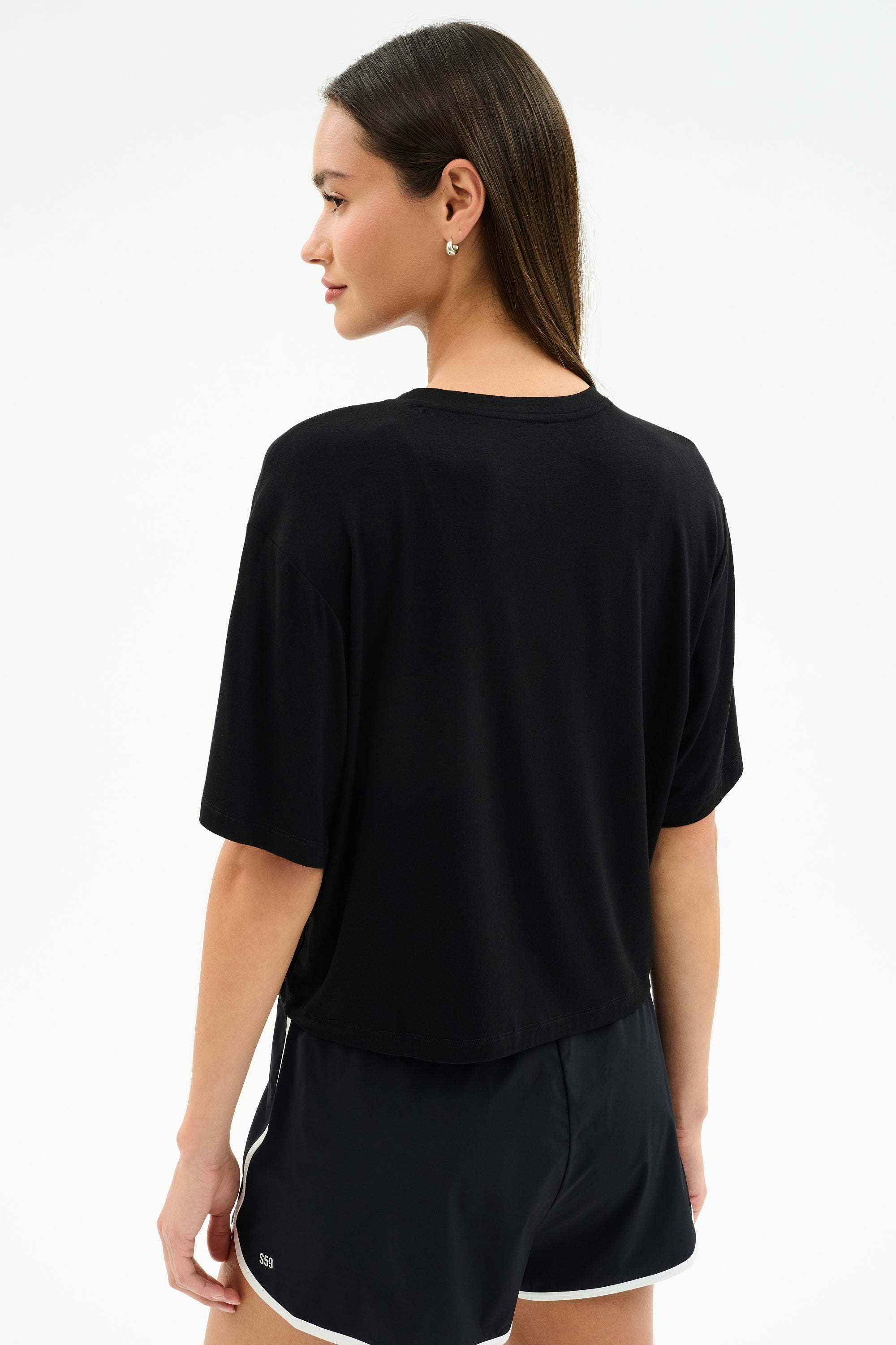 A woman faces away, wearing the SPLITS59 Djuna Oversized Jersey Tee in black with black athletic shorts featuring white trim against a light background—ideal gym workout attire.