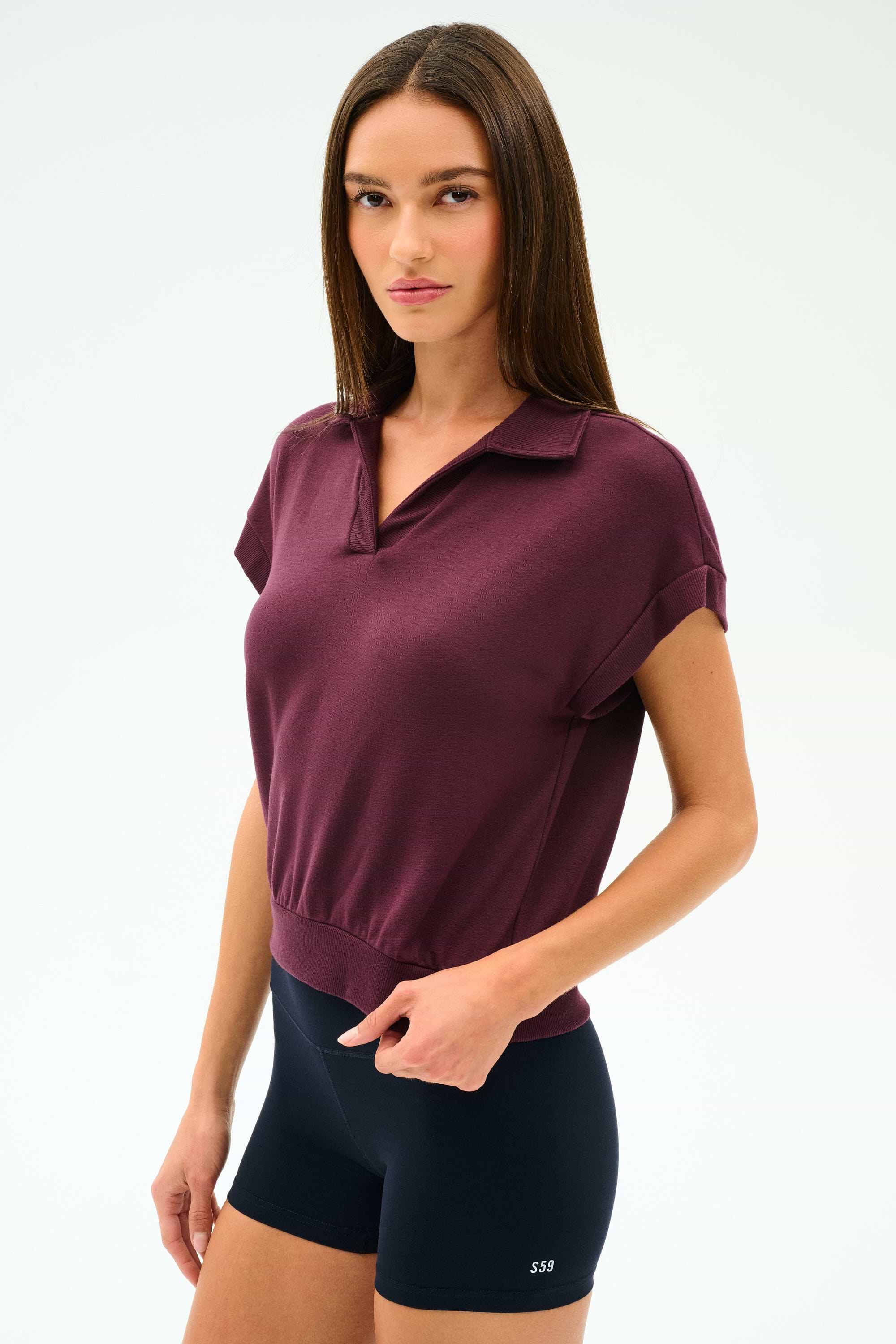A woman with straight brown hair wears the SPLITS59 Evie Fleece Sleeveless Polo in Dark Cherry and black fitted spandex shorts against a plain light background.