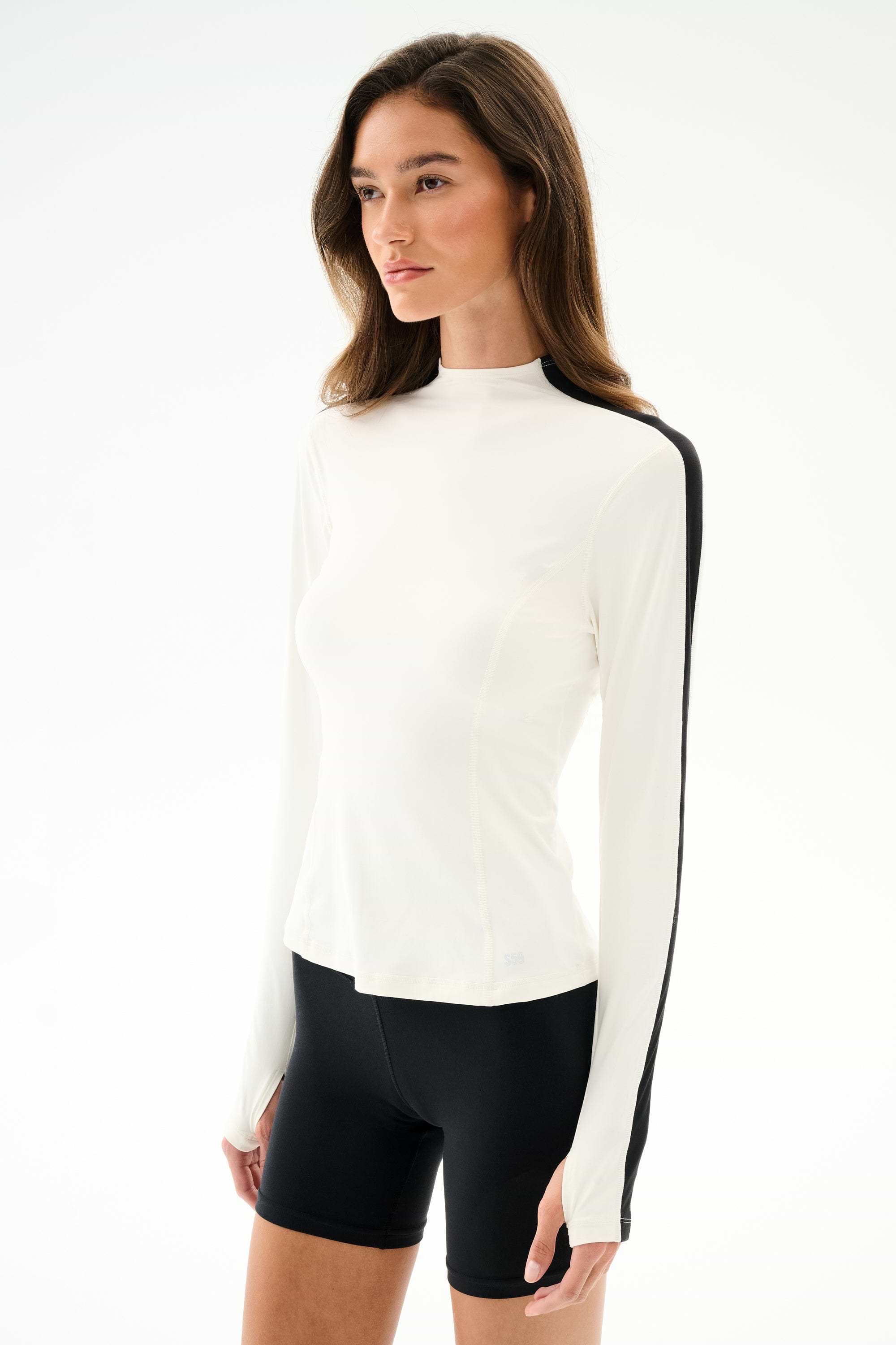 A woman wears the SPLITS59 Isabelle Airweight Lite Mock Neck - White/Black with black spandex shorts, standing against a plain white background.