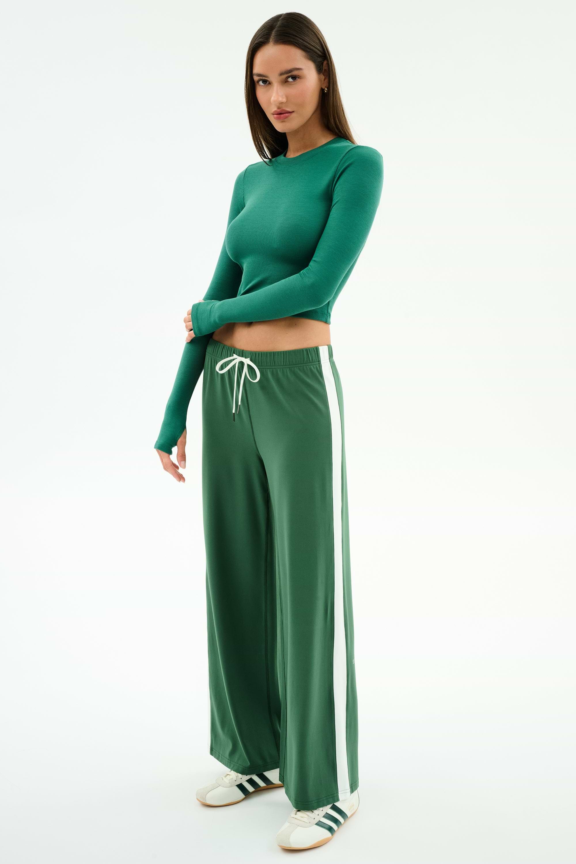 A woman wears the SPLITS59 Louise Rib Long Sleeve Crop in Arugula, paired with green wide-leg track pants featuring white side stripes and white sneakers with green accents, standing against a plain white background.