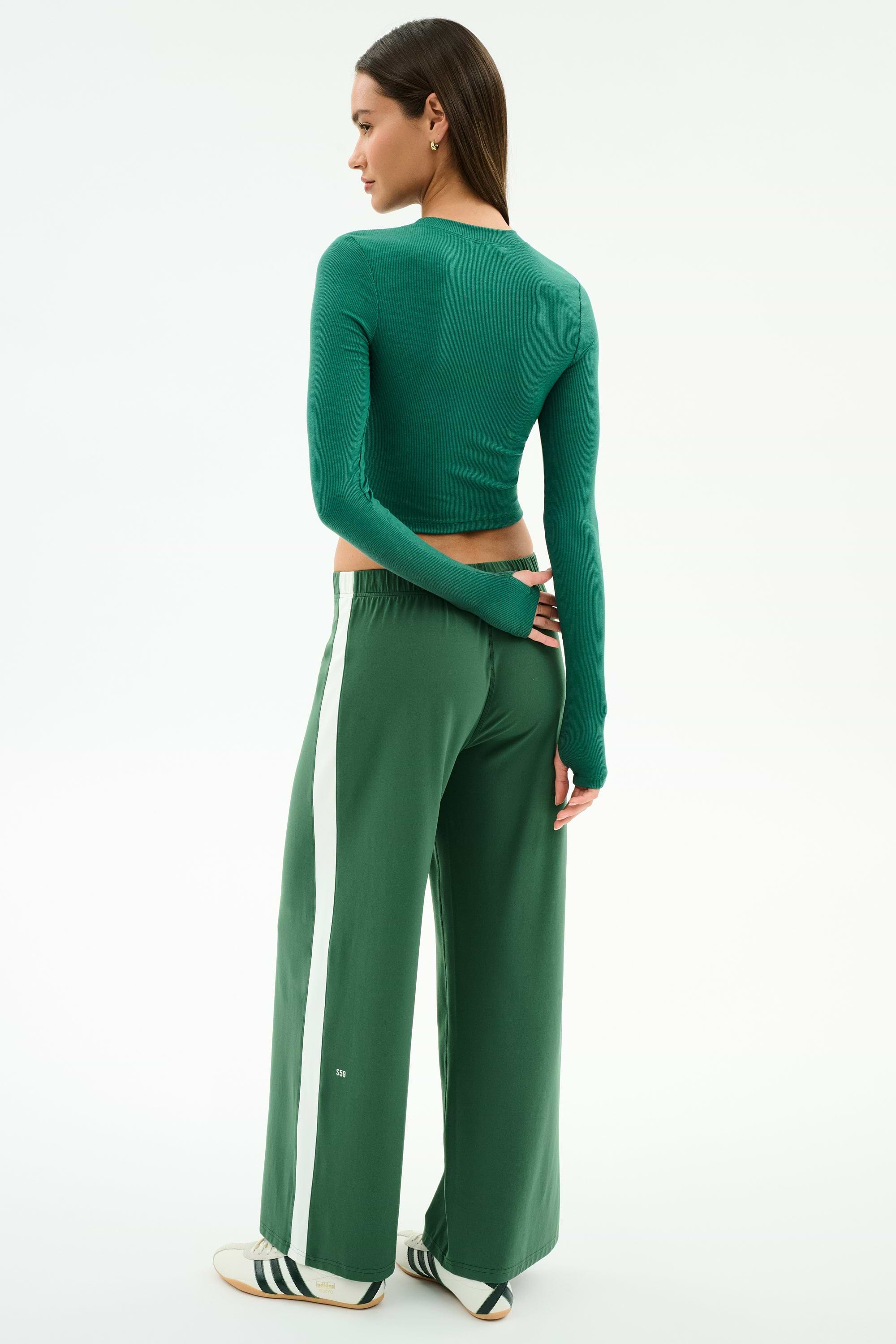 A woman stands facing away in the SPLITS59 Louise Rib Long Sleeve Crop in arugula and matching green wide-leg pants with a white stripe, paired with white sneakers featuring black stripes, set against a plain background.