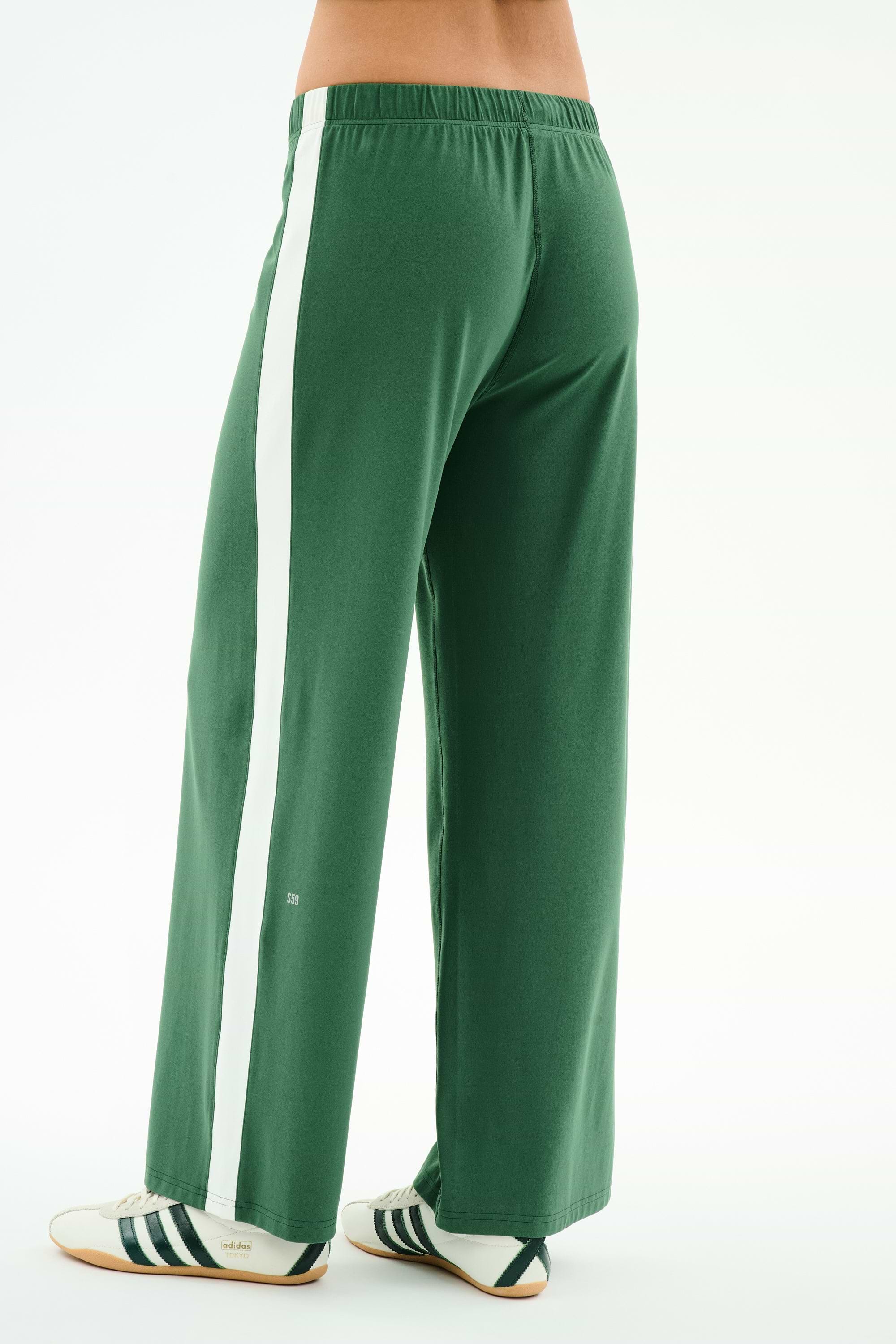 A person models SPLITS59’s Willow Airweight Wide Leg pants in Arugula/White with white sneakers featuring black stripes, standing against a white background.