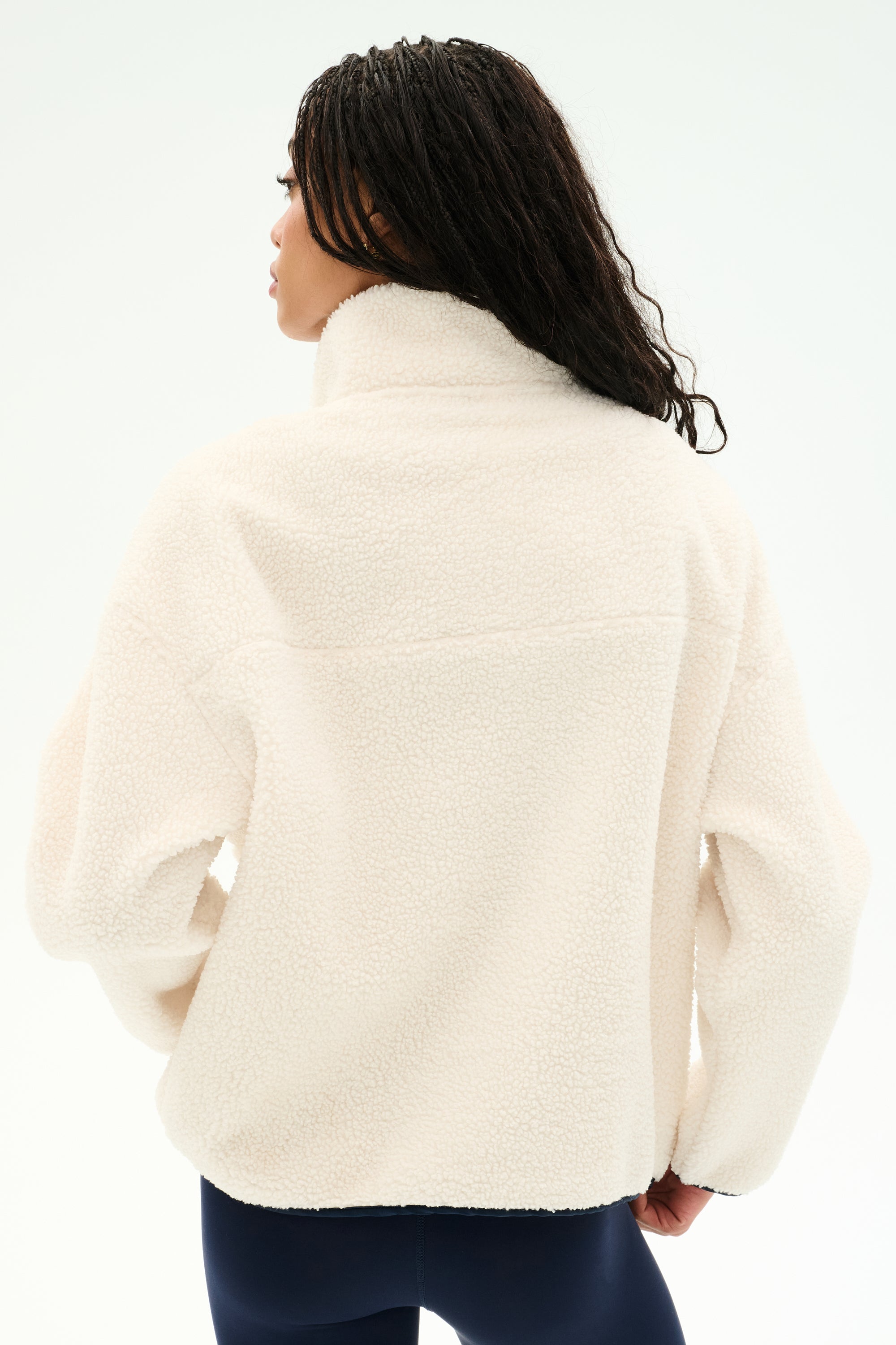A person with long braided hair models the SPLITS59 Reese Sherpa Half Zip Pullover in Creme/Indigo, crafted from soft 100% polyester, shown from the back and paired with dark leggings against a plain white background.