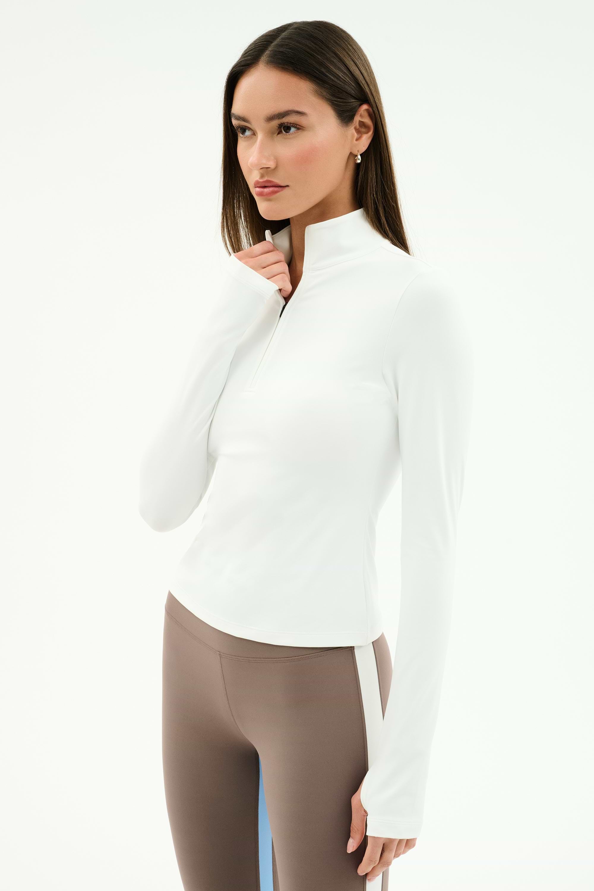A woman stands against a white background wearing the SPLITS59 Tess Rigor Half Zip Waist Length top in white paired with taupe leggings.