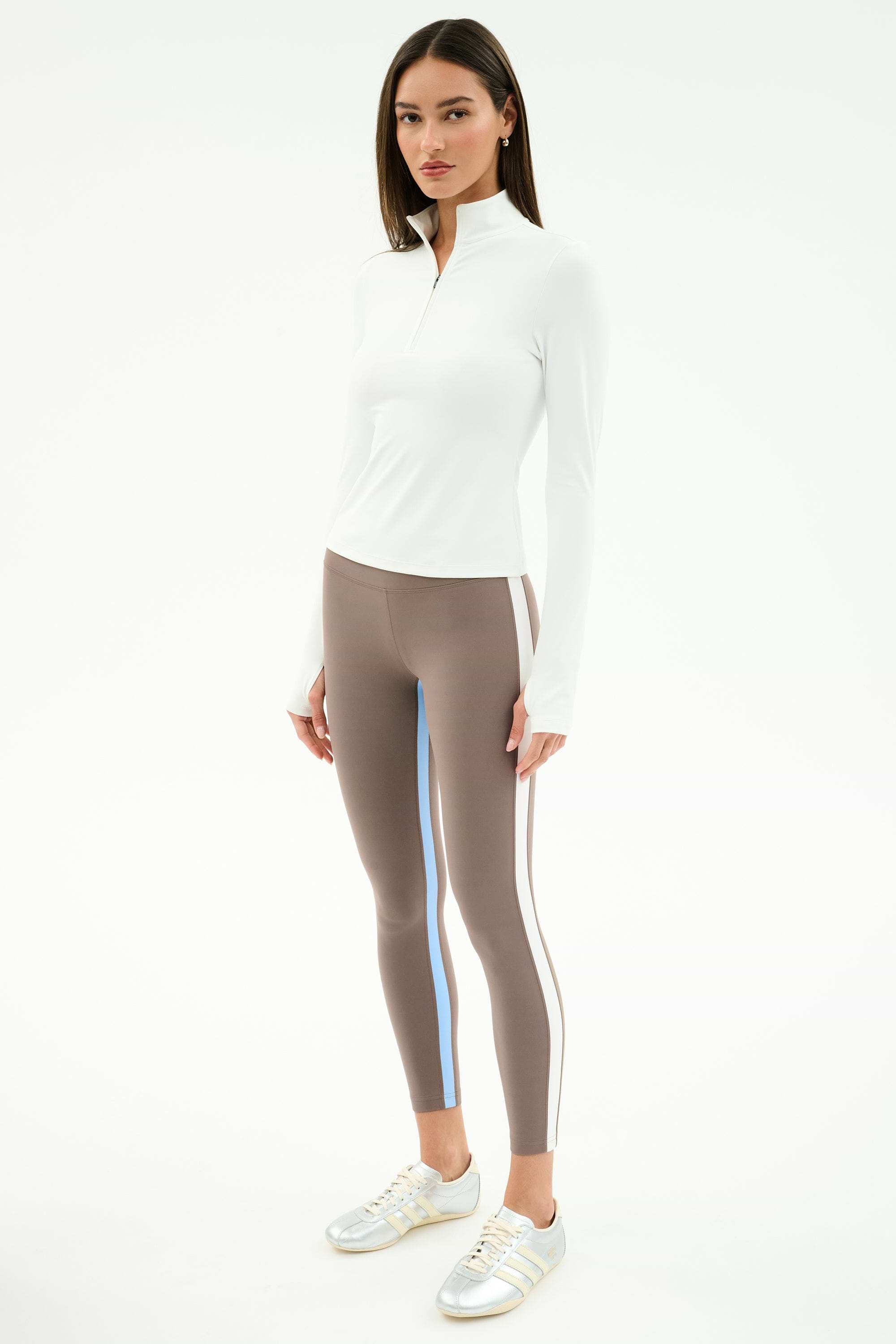 A woman wears the SPLITS59 Tess Rigor Half Zip Waist Length top in white, taupe leggings with blue and brown side stripes, and white sneakers, standing against a plain white background.
