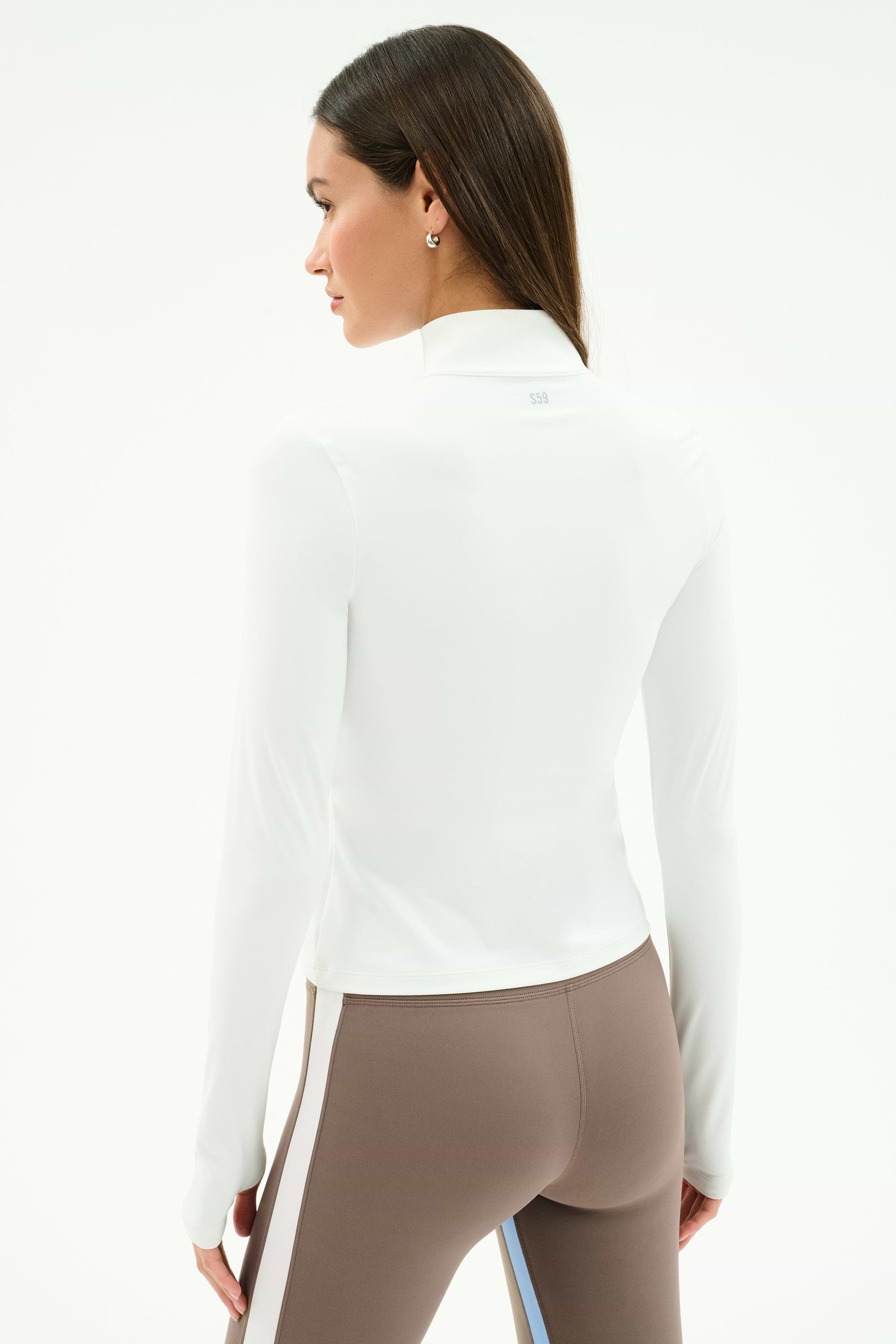 A woman faces away, wearing the SPLITS59 Tess Rigor Half Zip Waist Length top in white and high-waisted taupe leggings with white and blue side stripes made from stretchy nylon spandex.