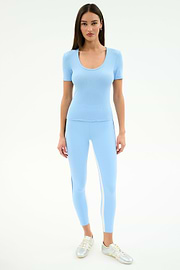 A woman stands facing forward in the SPLITS59 Ashby Rib Short Sleeve in Skylight with matching leggings featuring a side stripe, made from a soft modal-spandex blend, styled with silver sneakers against a plain white background.