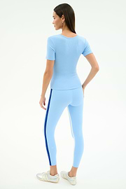 A woman faces away, wearing the SPLITS59 Ashby Rib Short Sleeve in Skylight and matching leggings with a dark blue side stripe, paired with white sneakers against a plain white background.