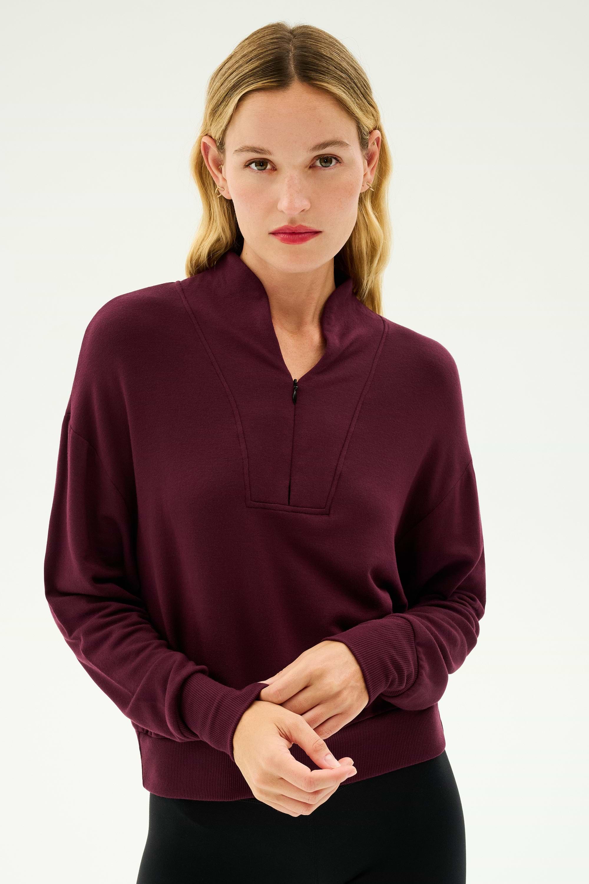 A person with long hair dressed in the cozy SPLITS59 Birdie Fleece Half Zip in Dark Cherry and black pants stands against a plain backdrop.