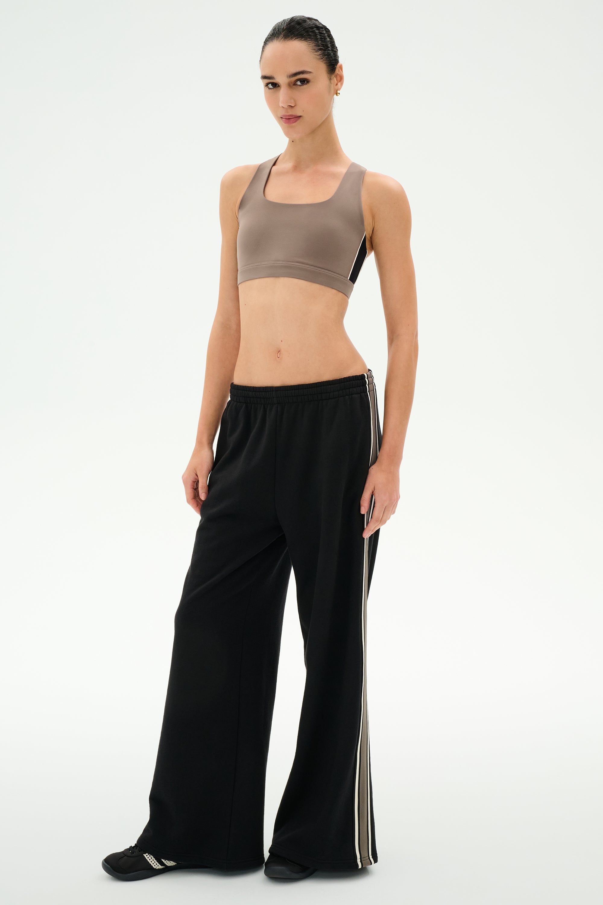 A person stands against a plain background, looking at the camera, wearing a taupe sports bra and the SPLITS59 Dallas French Terry Wide Leg Sweatpant in Black/Lentil—a pair of loose black pants with white side stripes crafted from soft cotton fabric.