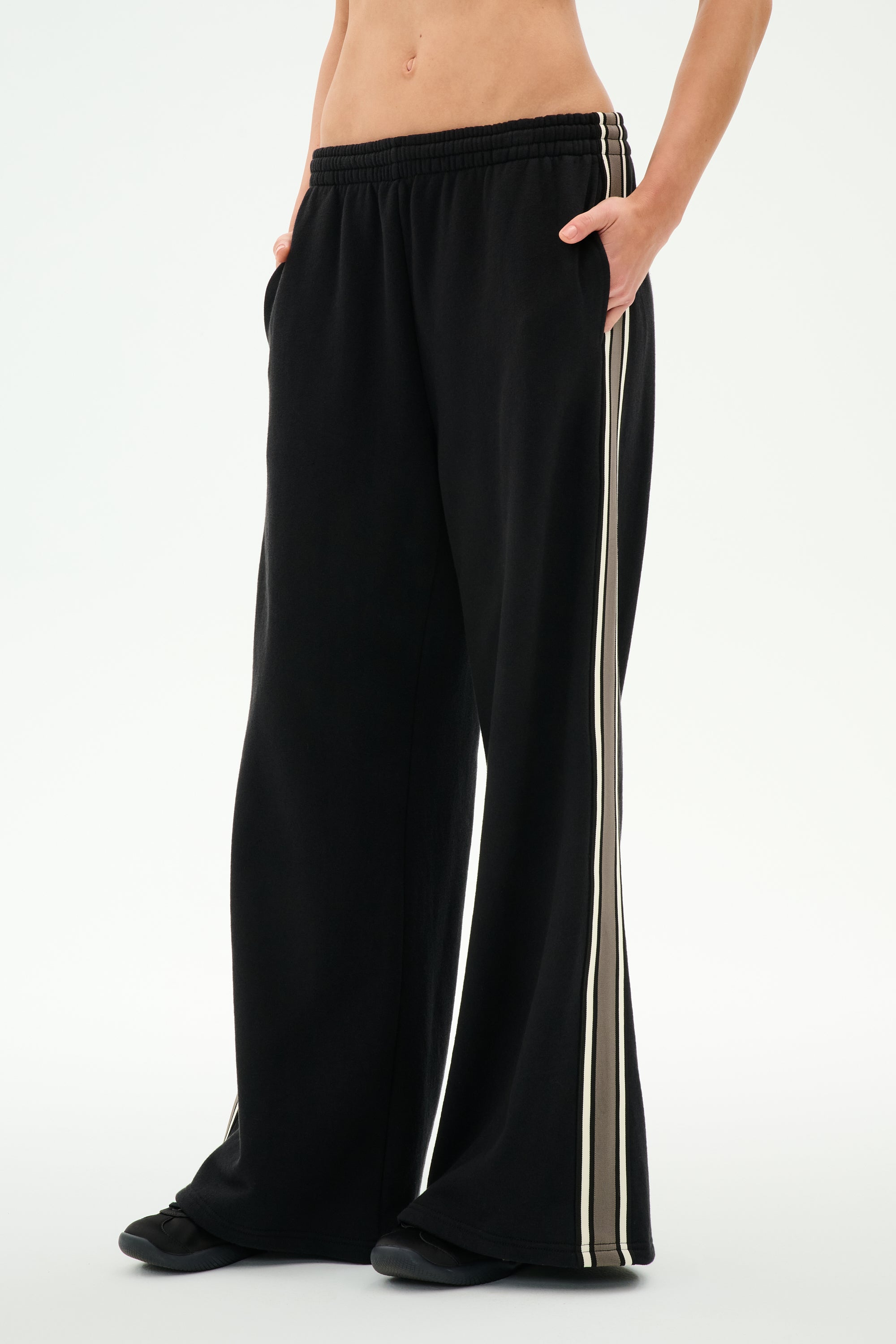 A person is wearing the SPLITS59 Dallas French Terry Wide Leg Sweatpant in Black/Lentil, featuring loose wide legs and white side stripes, crafted from soft 100% cotton, standing barefoot with hands in pockets against a plain light background; the image is cropped at the waist and ankles.