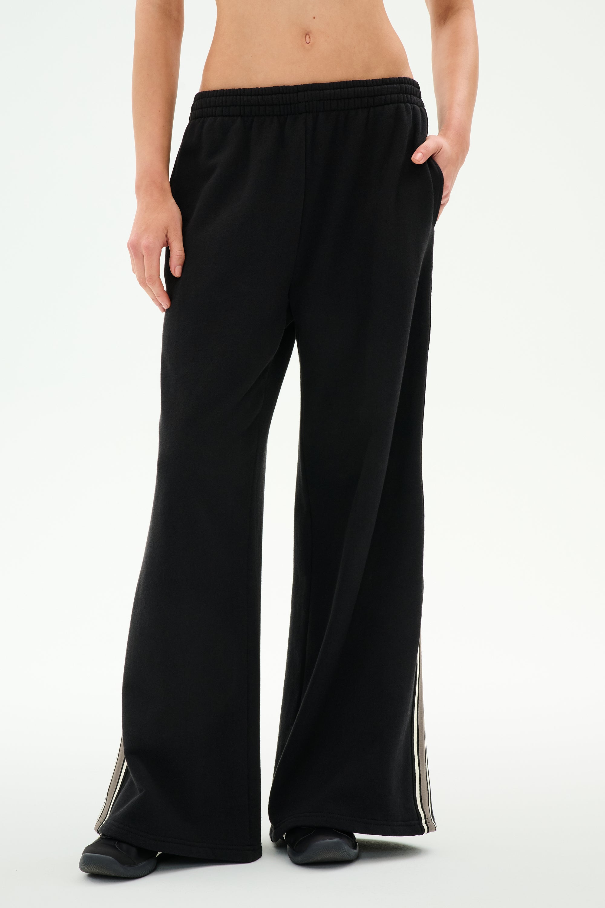 A person wearing the SPLITS59 Dallas French Terry Wide Leg Sweatpant in Black/Lentil, crafted from soft cotton fabric and featuring side stripes, stands with one hand in their pocket, shown from midriff to feet.