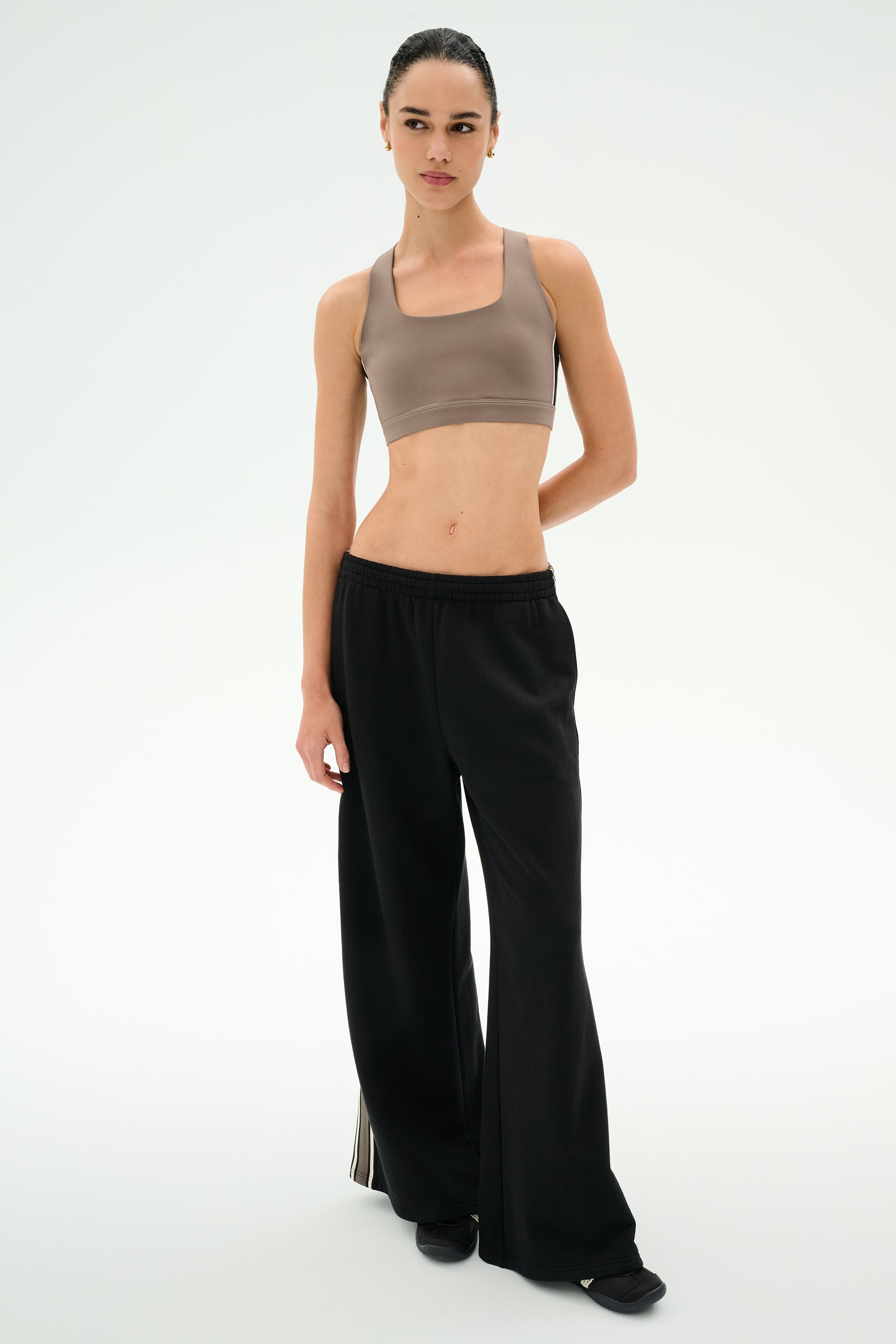 A person stands against a plain background, facing forward with a neutral expression, wearing the SPLITS59 Dallas French Terry Wide Leg Sweatpant in Black/Lentil—crafted from soft 100% cotton—which features loose black pants with side stripe detail and is paired with a taupe sports bra.