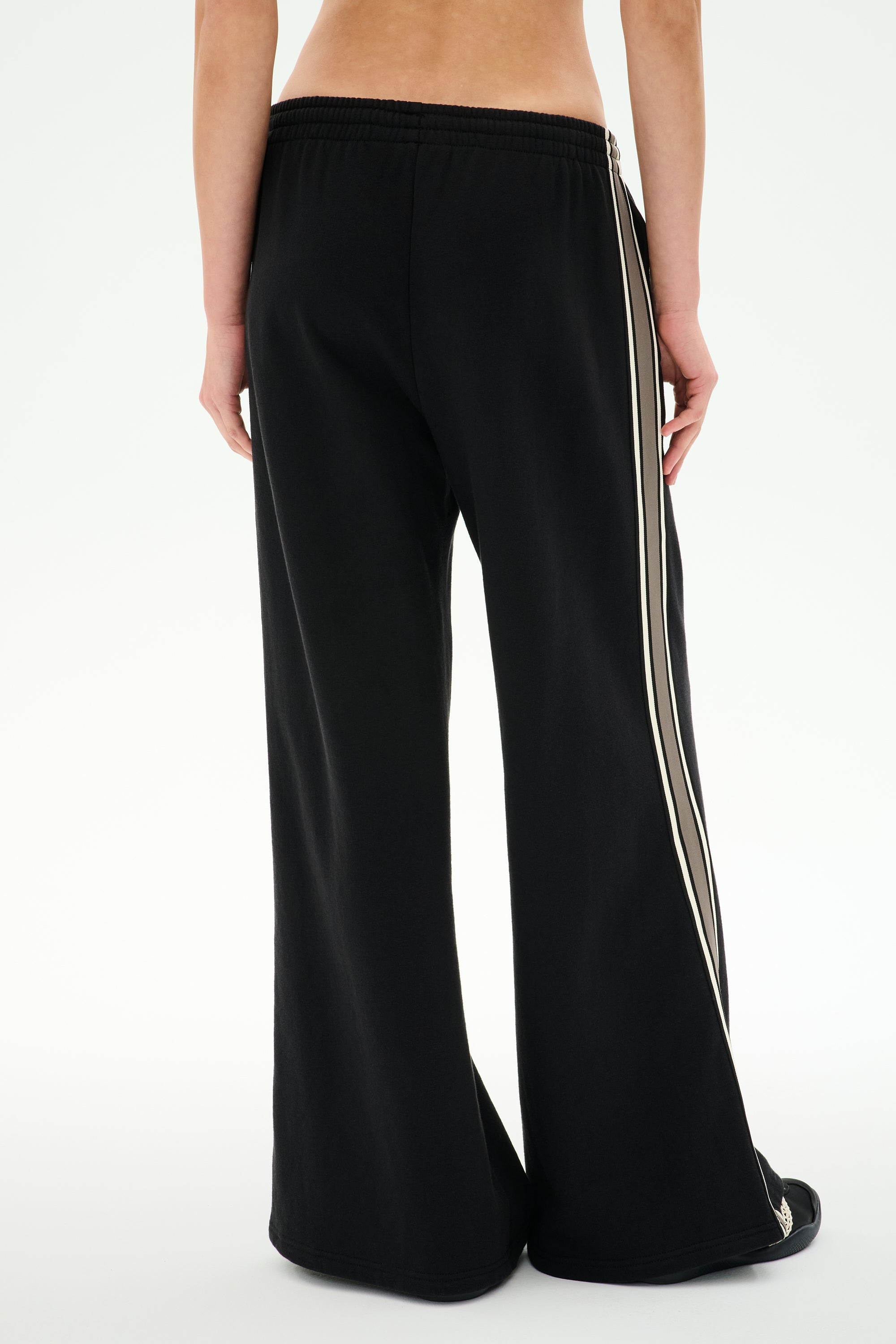 A person is shown from the back, wearing SPLITS59 Dallas French Terry Wide Leg Sweatpants in Black/Lentil—loose-fitting black pants with white side stripes made from soft cotton terry fabric—against a plain white background.