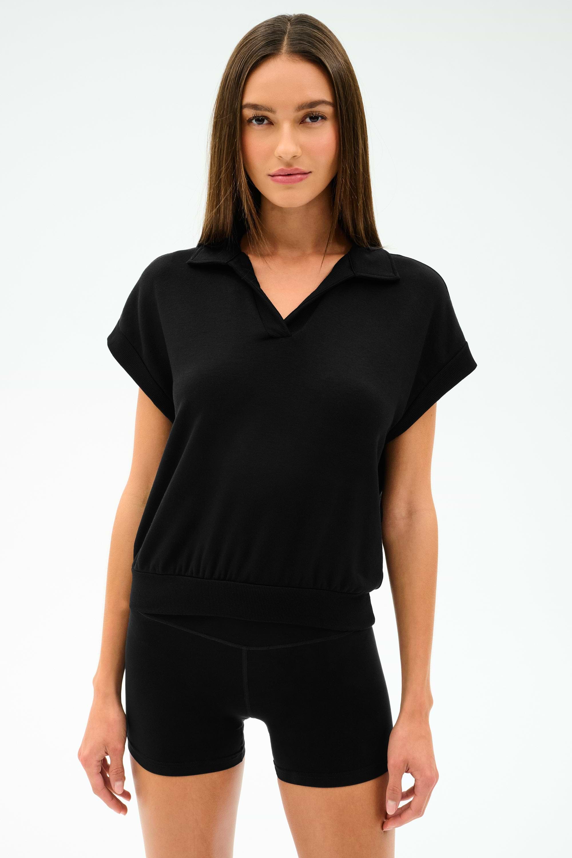 A woman wearing the SPLITS59 Evie Fleece Sleeveless Polo in black, paired with black shorts, stands against a plain light background. The look features sporty style and comfort.