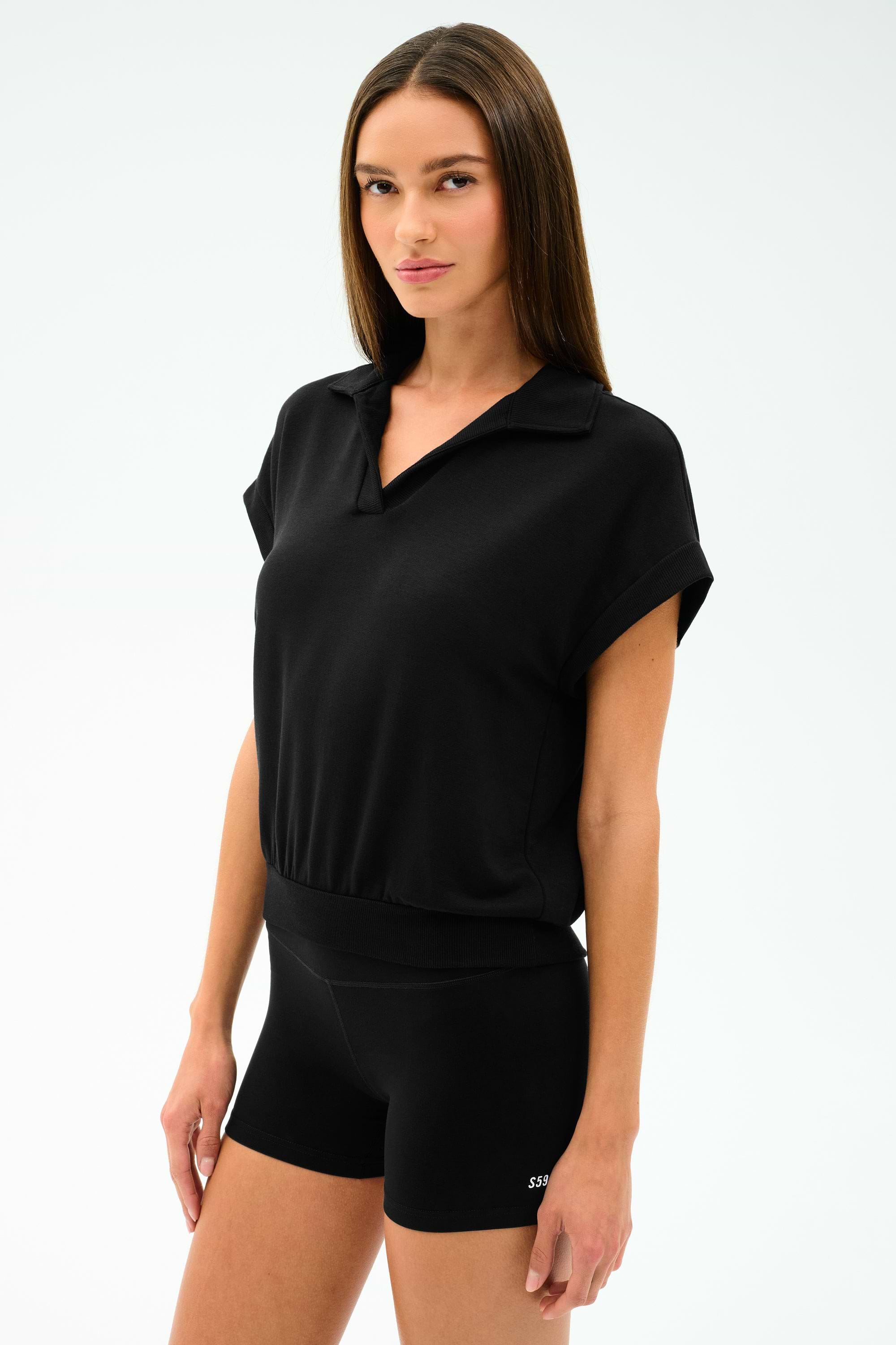 A woman with straight brown hair wears the SPLITS59 Evie Fleece Sleeveless Polo in black, paired with black fitted shorts, standing against a plain light background.