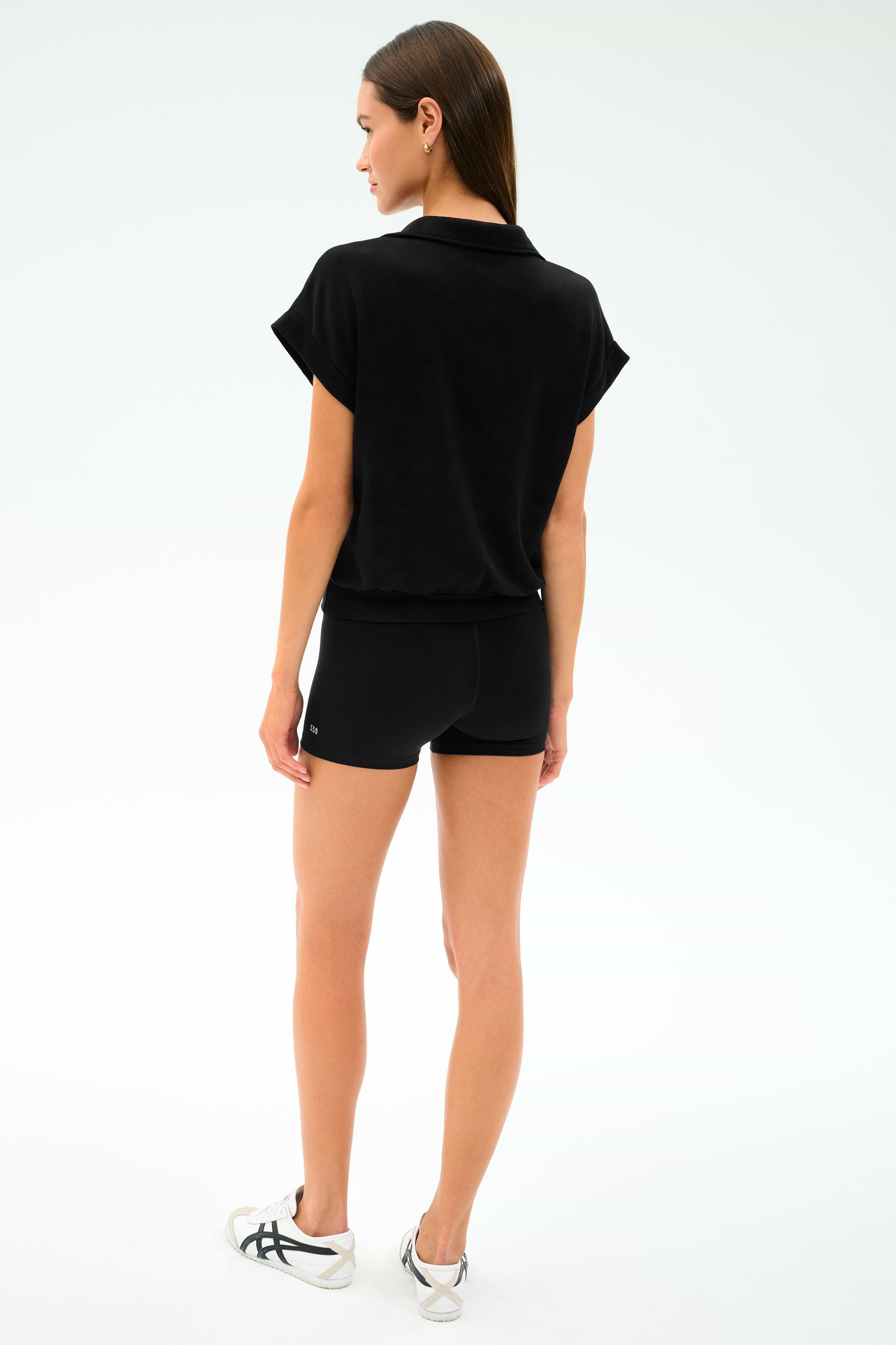 A woman faces away from the camera against a plain background, wearing the SPLITS59 Evie Fleece Sleeveless Polo in black paired with black shorts and white sneakers.