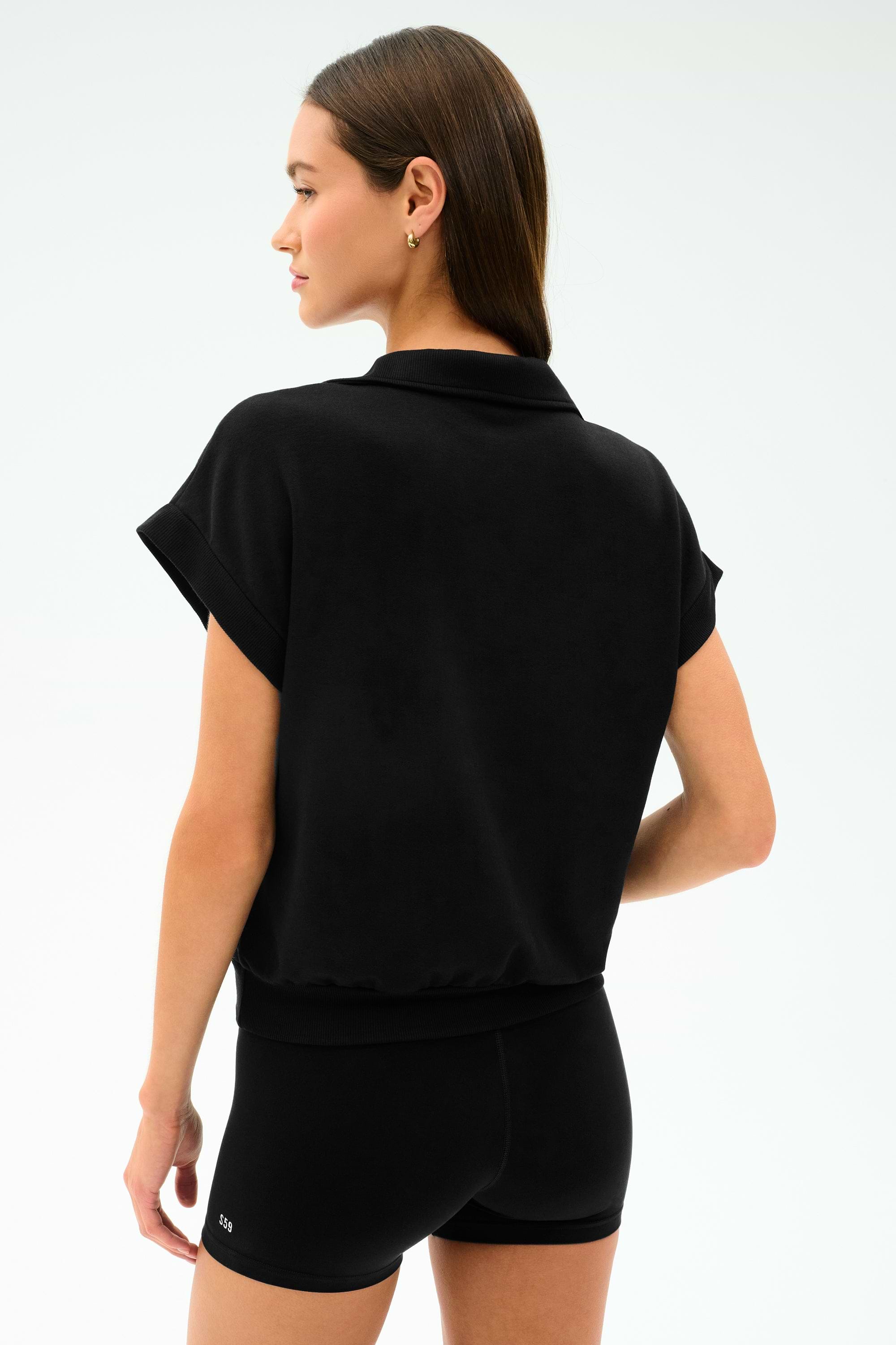 A woman faces away, wearing the SPLITS59 Evie Fleece Sleeveless Polo in black, paired with matching shorts, set against a plain light background.