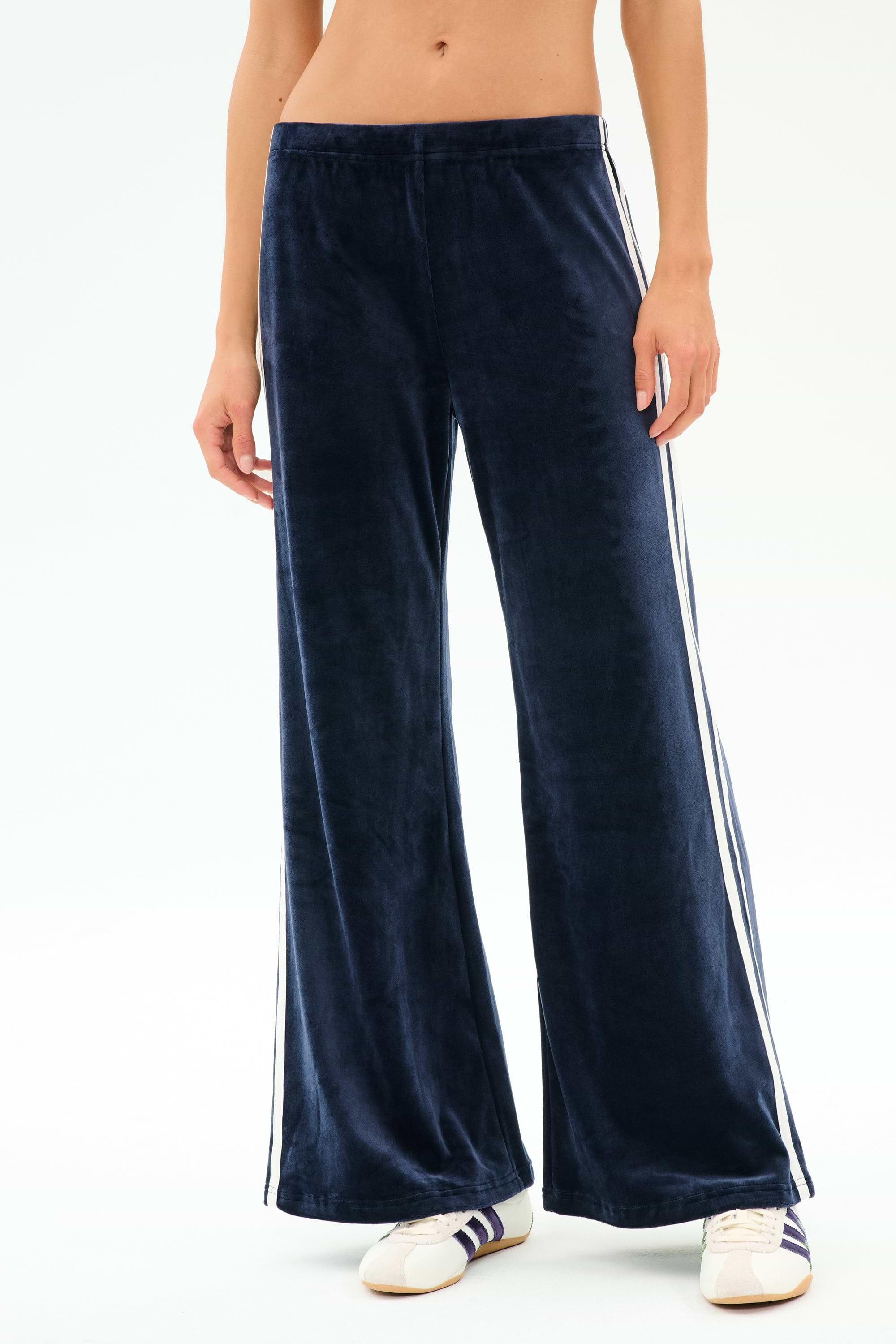 A person wears the SPLITS59 Heritage Velour Pant in Indigo/White, made with spandex and polyester, featuring white side stripes, paired with white athletic shoes against a plain light background.
