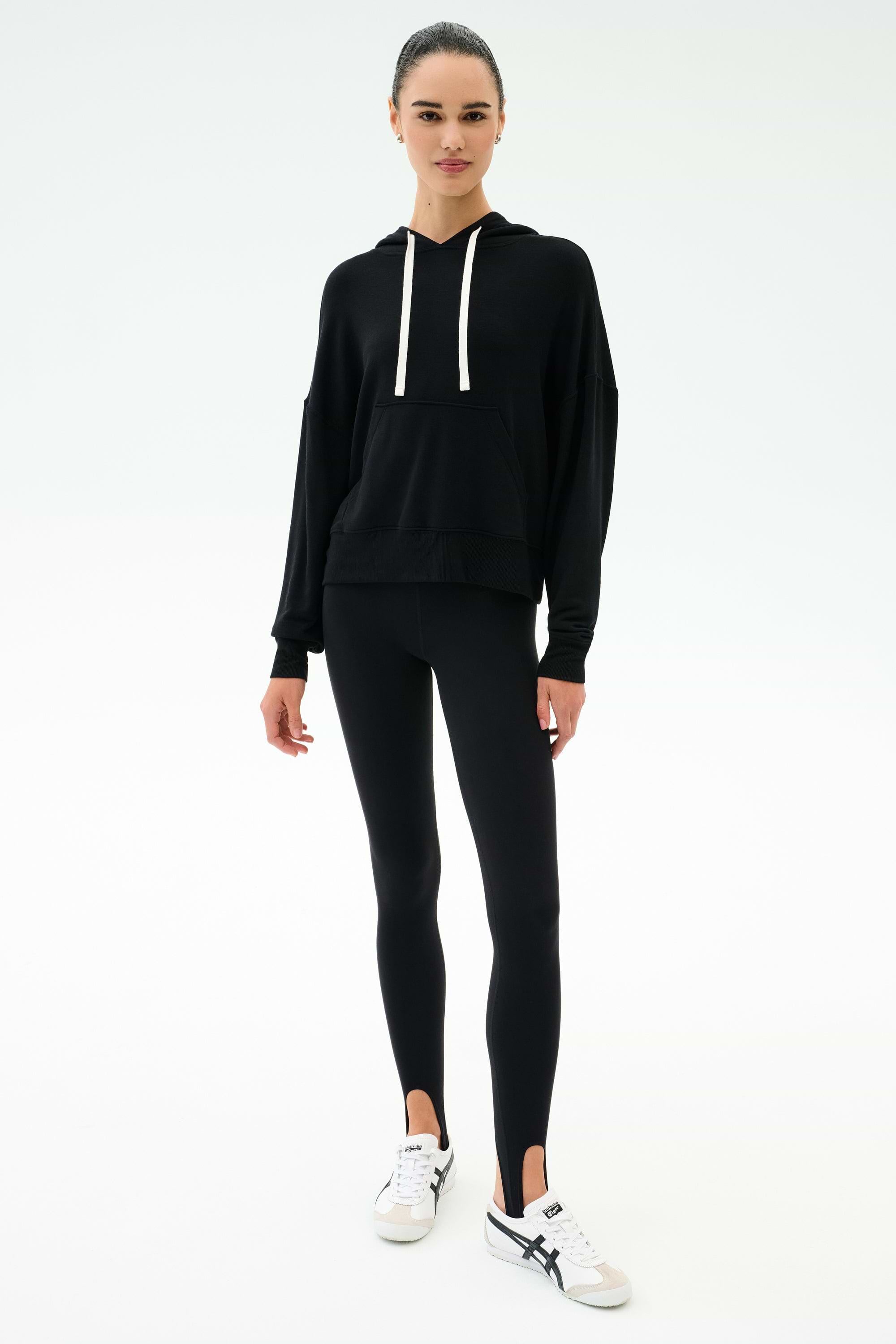 A person stands facing forward in a SPLITS59 Joey Fleece Hoodie in black, black spandex blend leggings with stirrups, and white sneakers against a plain white background.