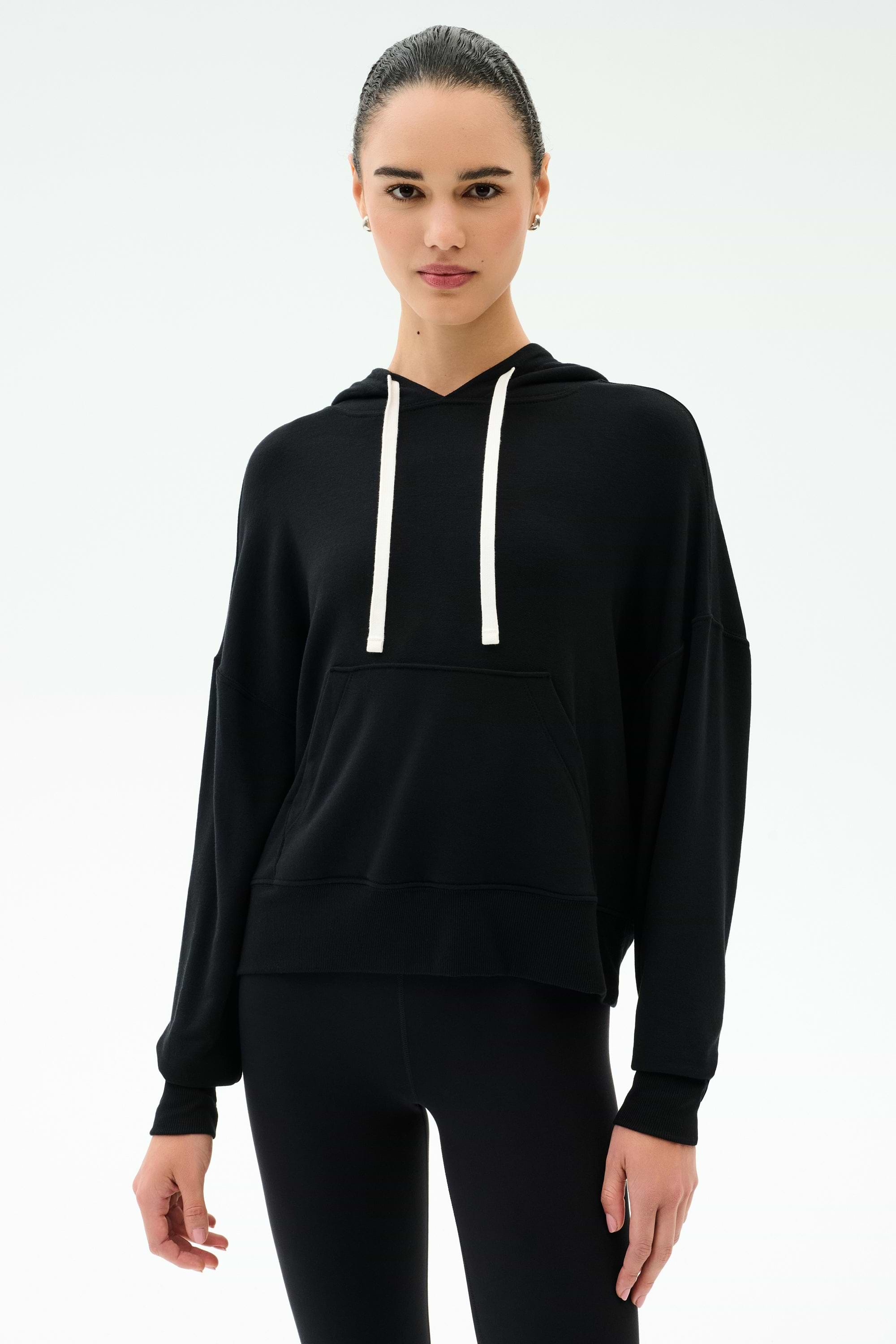 A person wears the SPLITS59 Joey Fleece Hoodie in black with white drawstrings and black spandex-blend leggings, standing against a plain white background.