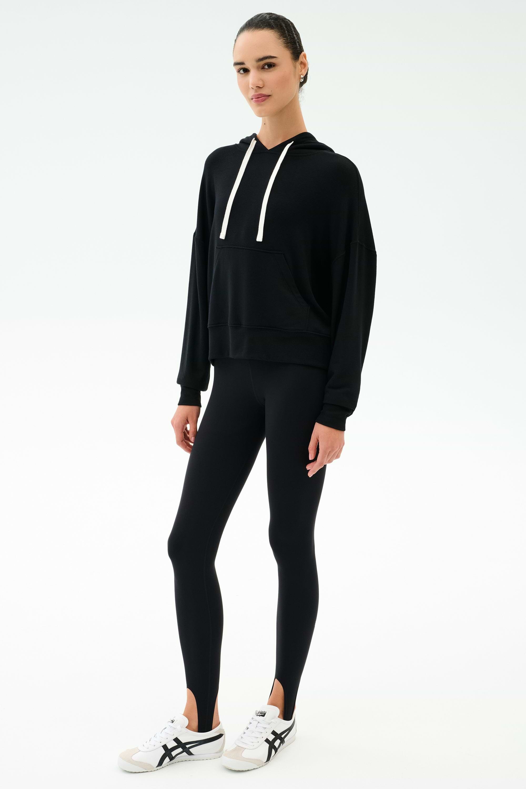 A person stands facing forward against a plain white background, wearing the SPLITS59 Joey Fleece Hoodie in black, black leggings, and white sneakers with black accents.