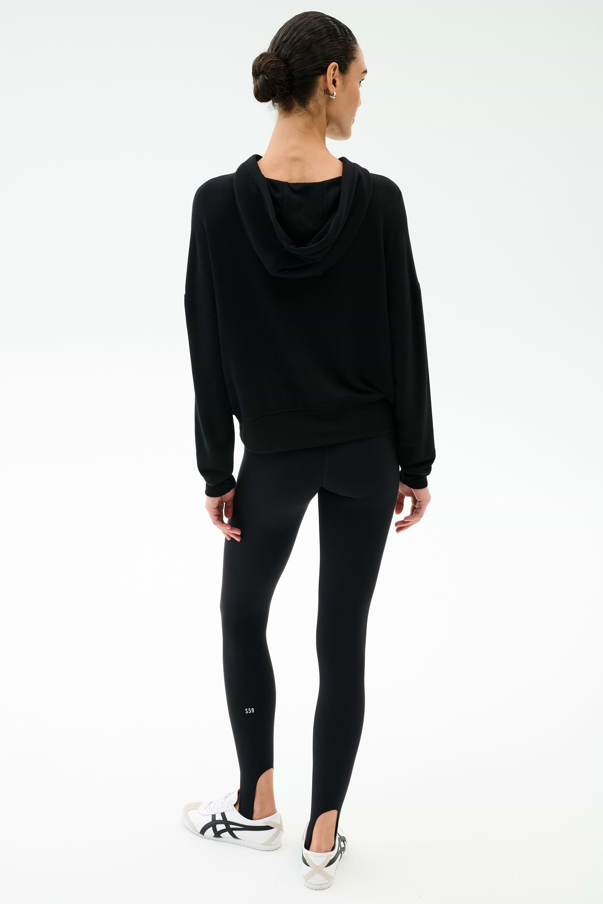 A woman stands with her back to the camera, wearing the SPLITS59 Joey Fleece Hoodie in black and matching leggings, paired with white sneakers, against a plain white background.