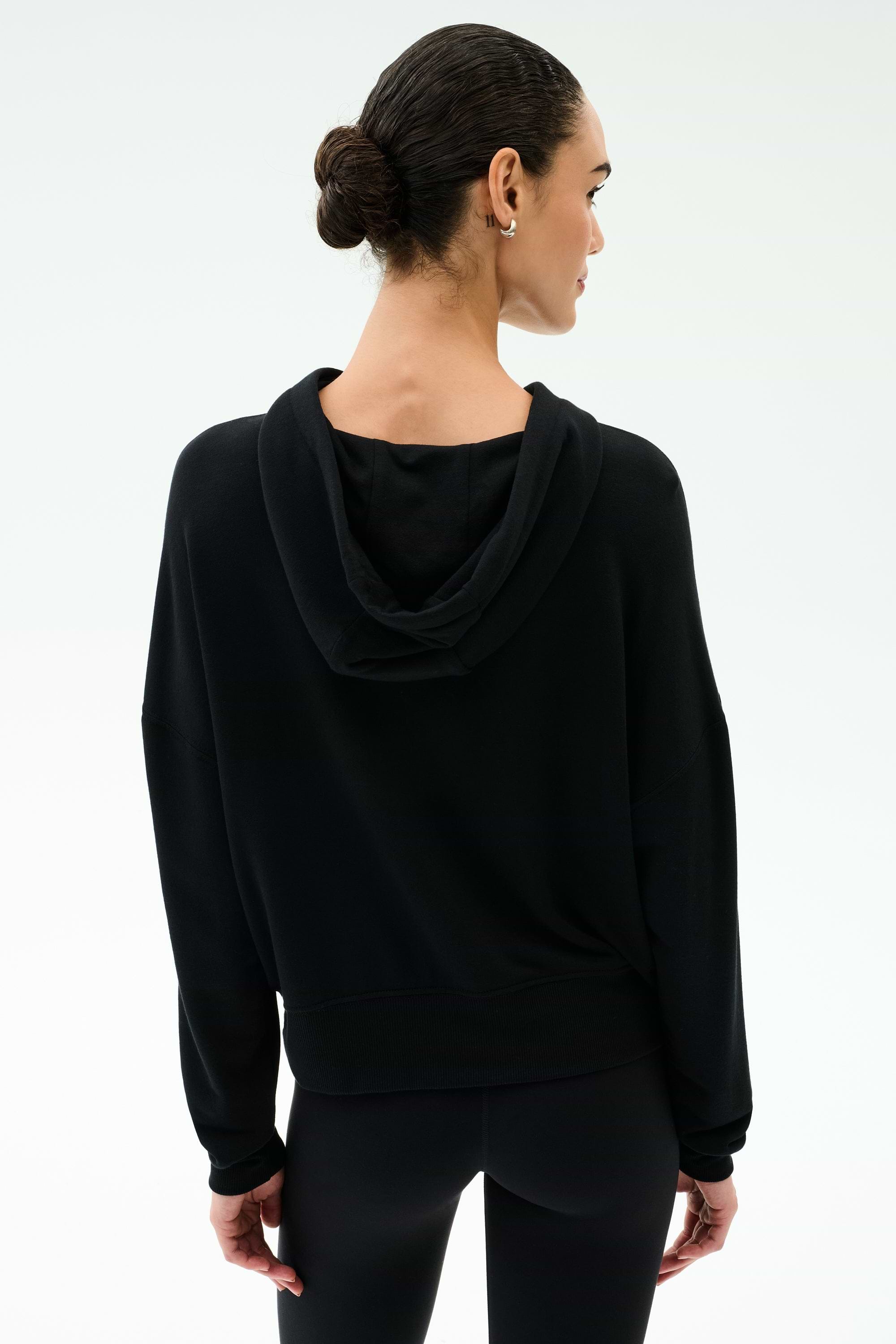 A woman with dark hair in a low bun is seen from behind wearing the SPLITS59 Joey Fleece Hoodie in black against a plain light background.