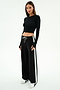 A woman stands facing slightly to the side, wearing the SPLITS59 Louise Rib Long Sleeve Crop in black, paired with black wide-leg pants featuring white side stripes and white sneakers, against a plain white background.