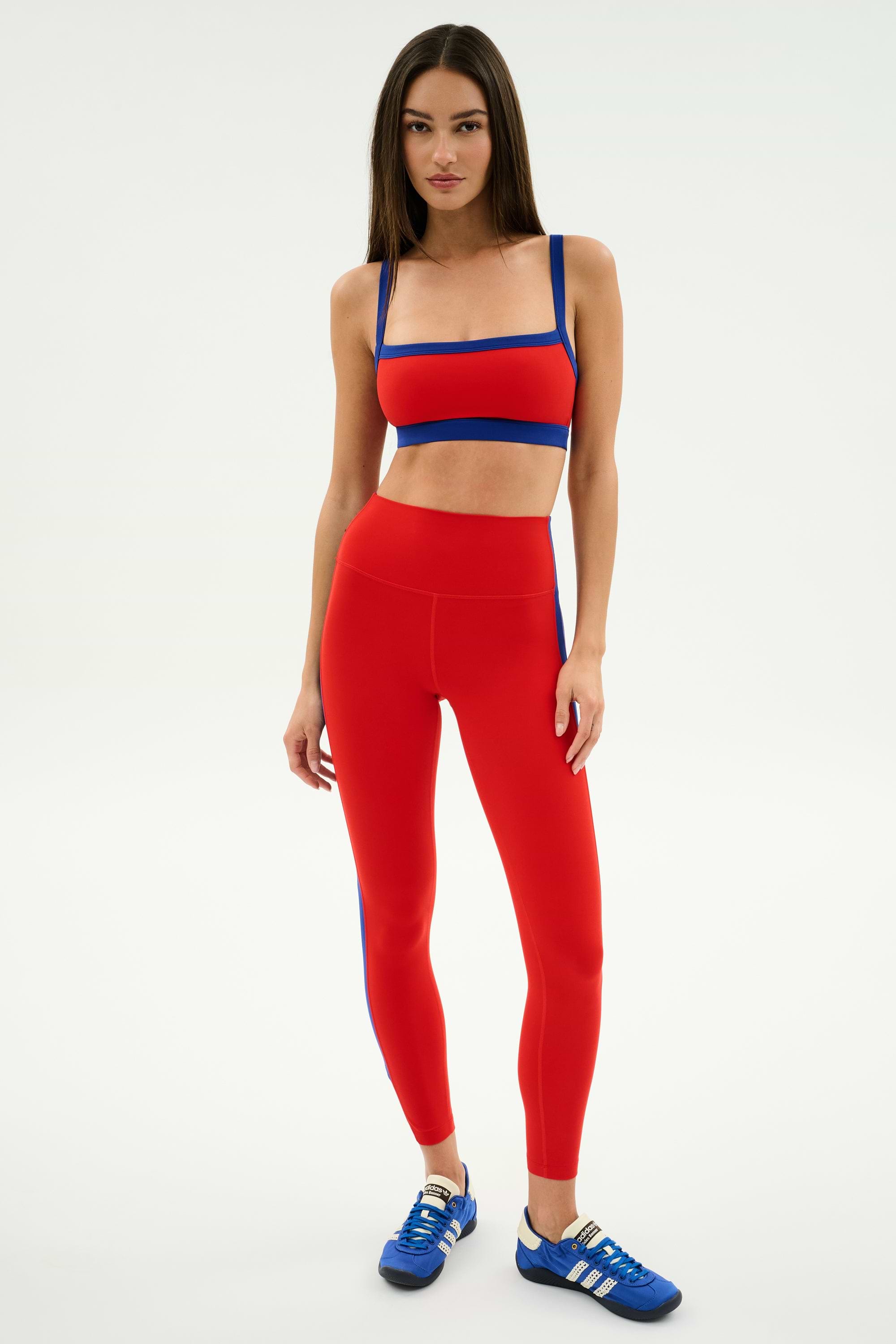 A woman stands against a plain background wearing the SPLITS59 Monah Rigor Bra in Lotus Red/Cobalt, paired with matching leggings and blue athletic shoes.