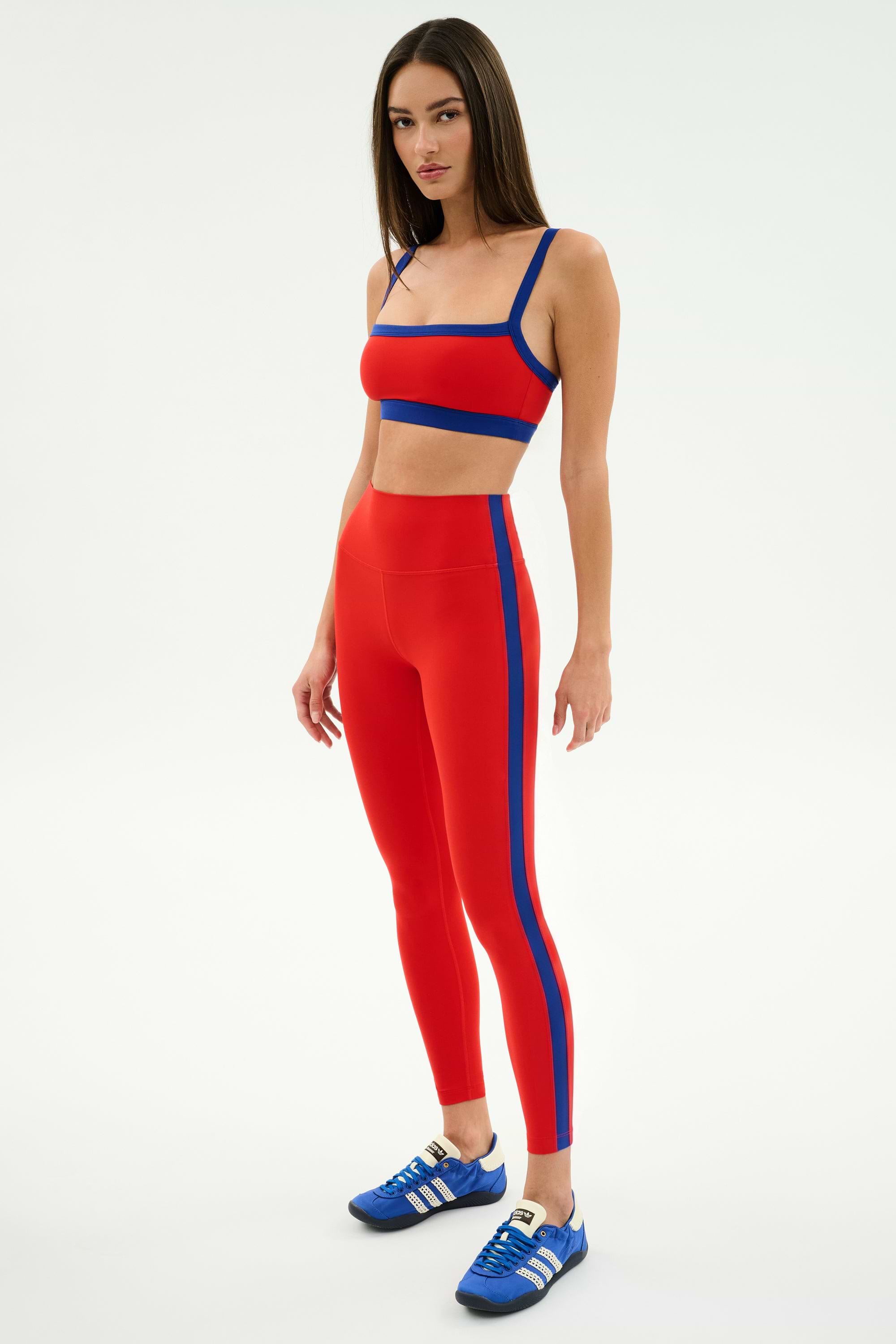 A woman models the SPLITS59 Monah Rigor Bra in Lotus Red/Cobalt, paired with matching high-waisted leggings and blue athletic shoes, standing confidently against a plain white background.
