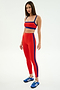 A woman models the SPLITS59 Monah Rigor Bra in Lotus Red/Cobalt, paired with matching high-waisted leggings and blue athletic shoes, standing confidently against a plain white background.