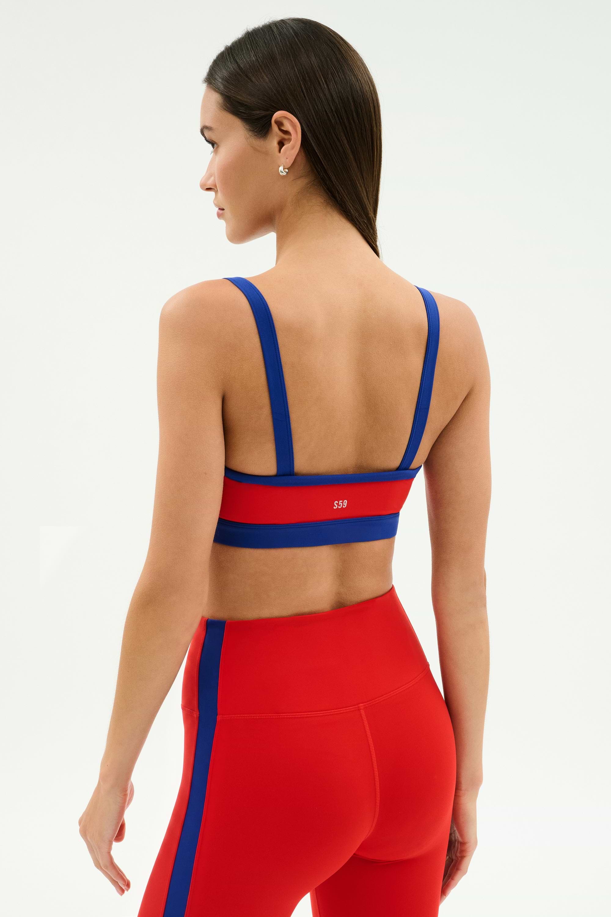 A woman wearing a SPLITS59 Monah Rigor Bra in Lotus Red/Cobalt with matching high-waisted leggings stands with her back to the camera against a plain background.