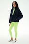 A woman stands against a plain white background wearing the SPLITS59 Reese Sherpa Half Zip Pullover in Indigo/Limeade, featuring neon green accents. The pullover is crafted from soft polyester material for comfort and durability. She pairs it with neon green leggings and white athletic sneakers.