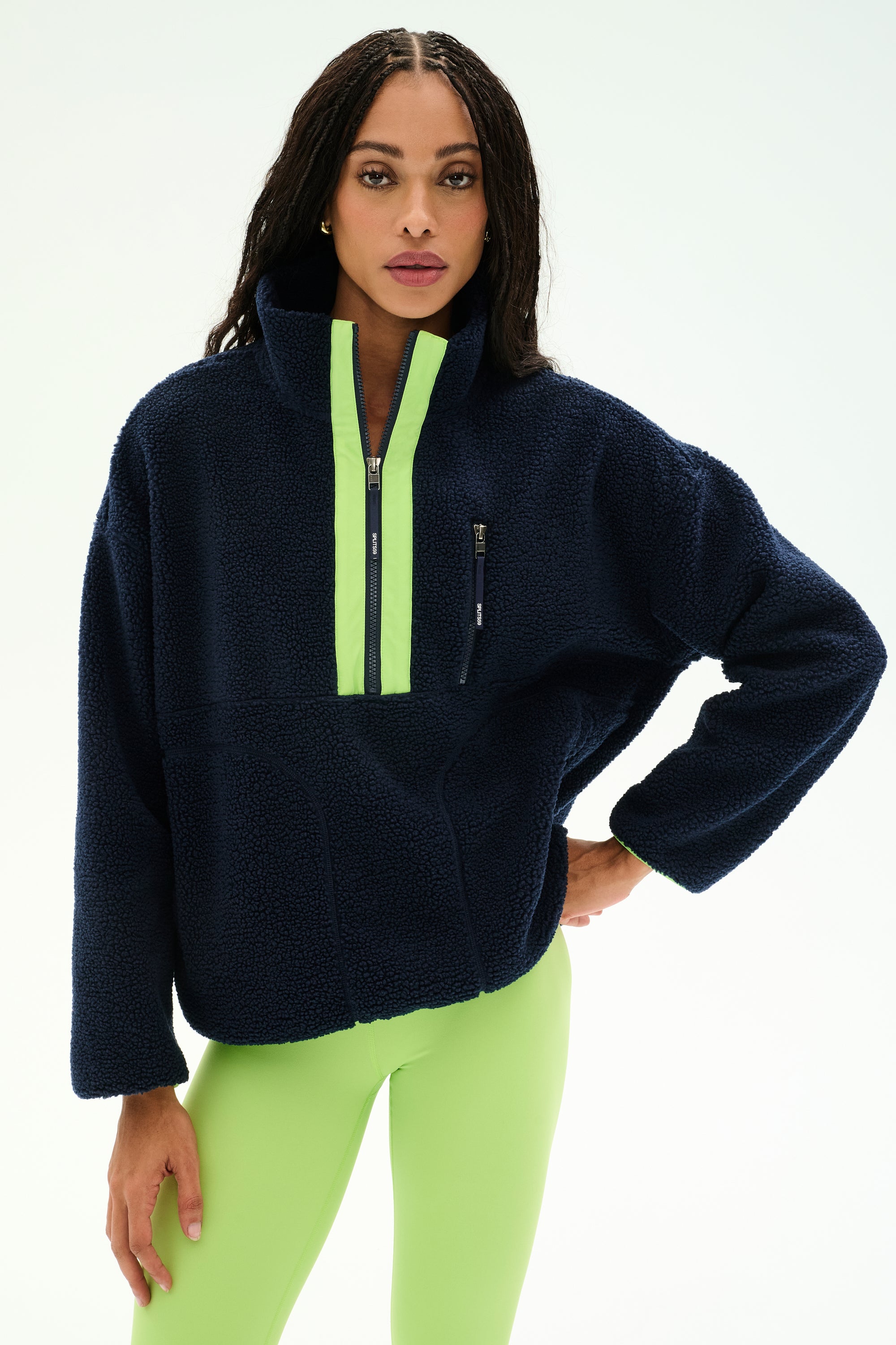 A woman stands against a plain light background wearing the SPLITS59 Reese Sherpa Half Zip Pullover in Indigo/Limeade, featuring soft polyester fabric and neon green accents, paired with neon green leggings. She poses with one hand on her hip.