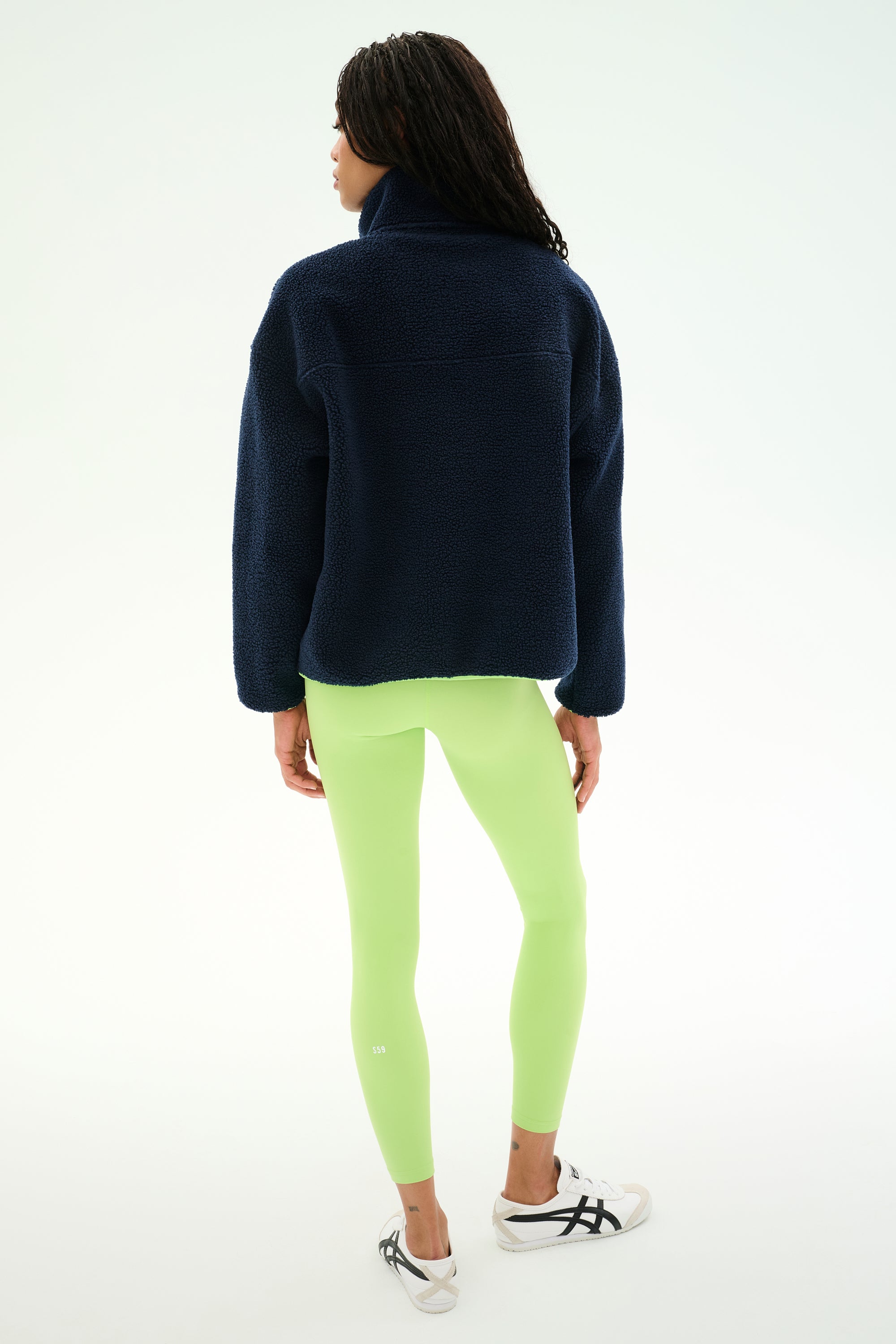 A woman stands with her back to the camera, wearing the SPLITS59 Reese Sherpa Half Zip Pullover in Indigo/Limeade, paired with neon green leggings and white sneakers against a plain light background.