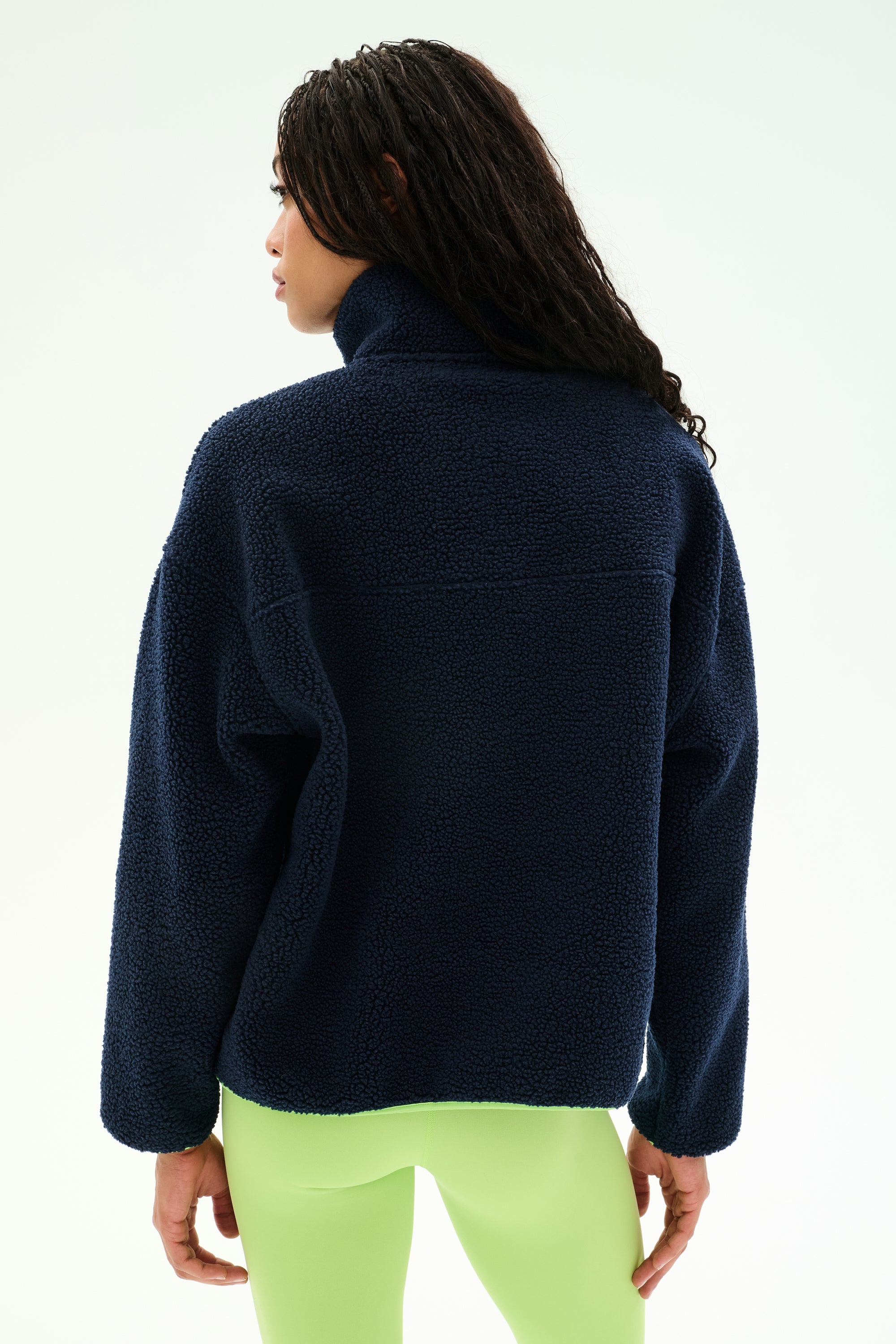 A person stands with their back to the camera, wearing the SPLITS59 Reese Sherpa Half Zip Pullover in Indigo/Limeade, paired with light green leggings, against a plain background.
