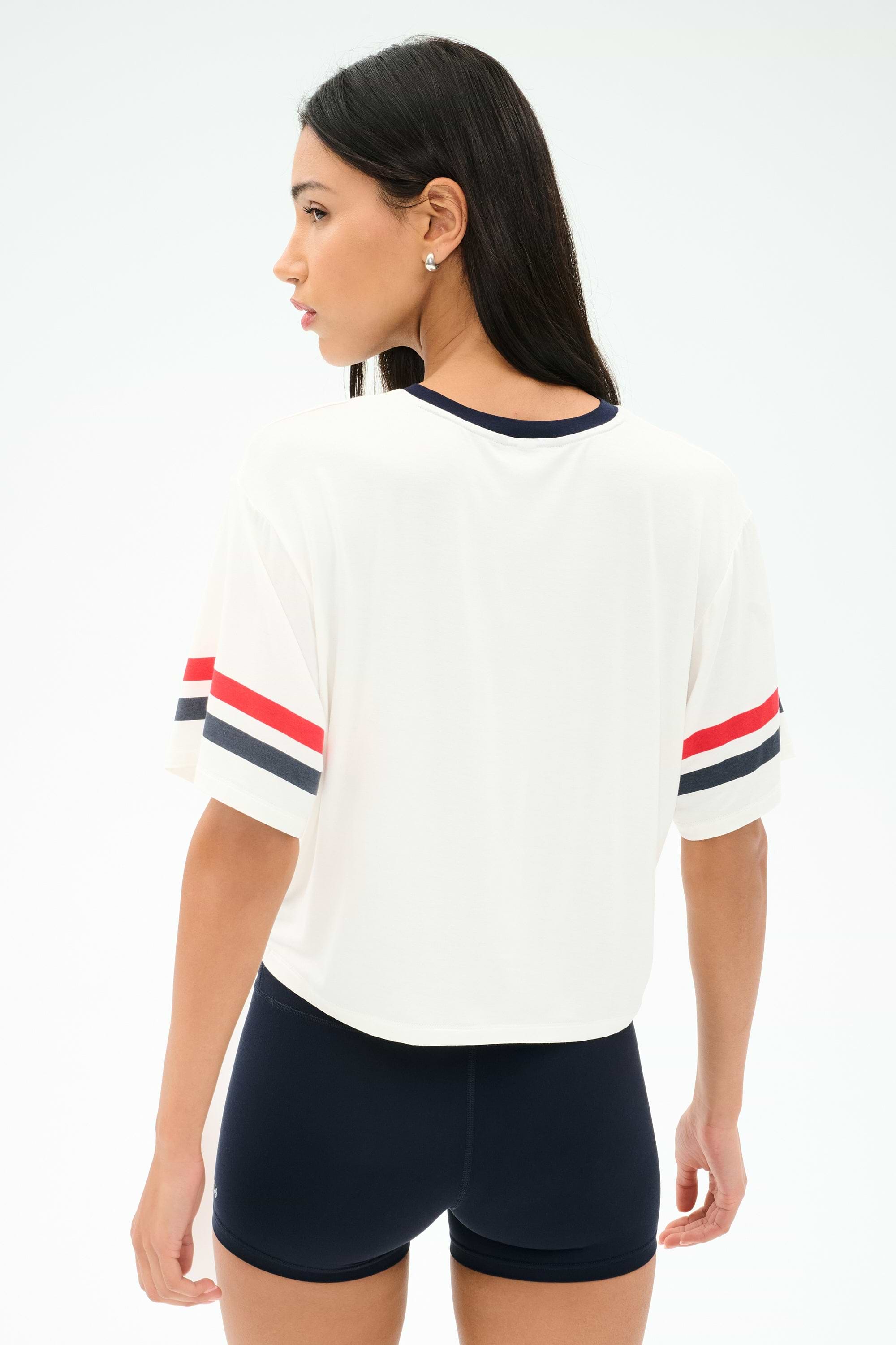 A woman stands with her back to the camera, wearing the SPLITS59 Rex Varsity Jersey V-Neck in White/59 with red and navy sleeve stripes, paired with navy shorts. The outfit is crafted from soft viscose for comfort against a plain light background.