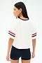 A woman stands with her back to the camera, wearing the SPLITS59 Rex Varsity Jersey V-Neck in White/59 with red and navy sleeve stripes, paired with navy shorts. The outfit is crafted from soft viscose for comfort against a plain light background.