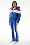A woman wears the SPLITS59 Aspen Puffer Jacket - Cobalt/Creme with blue flared pants, side stripes, and silver sneakers against a plain white background.