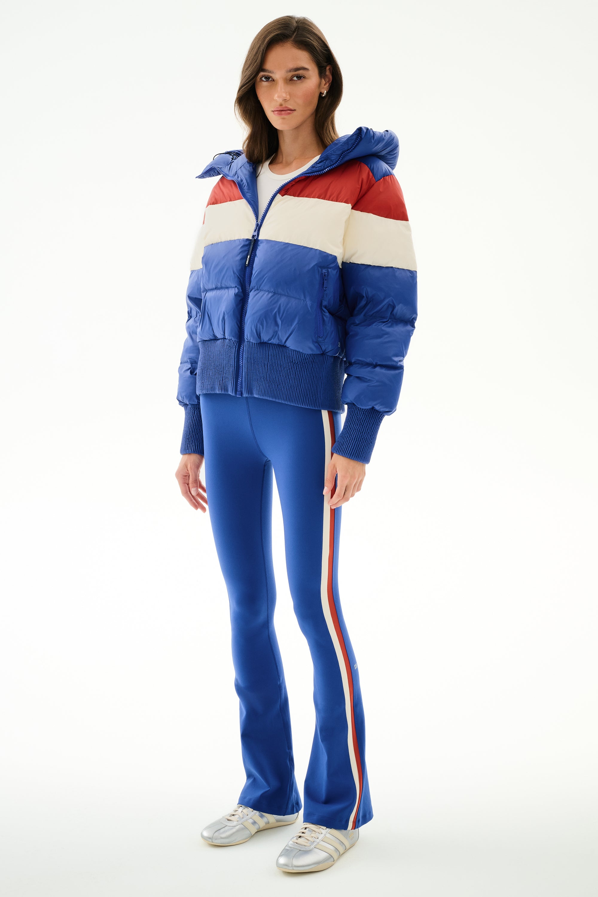 A woman wears the SPLITS59 Aspen Puffer Jacket - Cobalt/Creme with blue flared pants, side stripes, and silver sneakers against a plain white background.